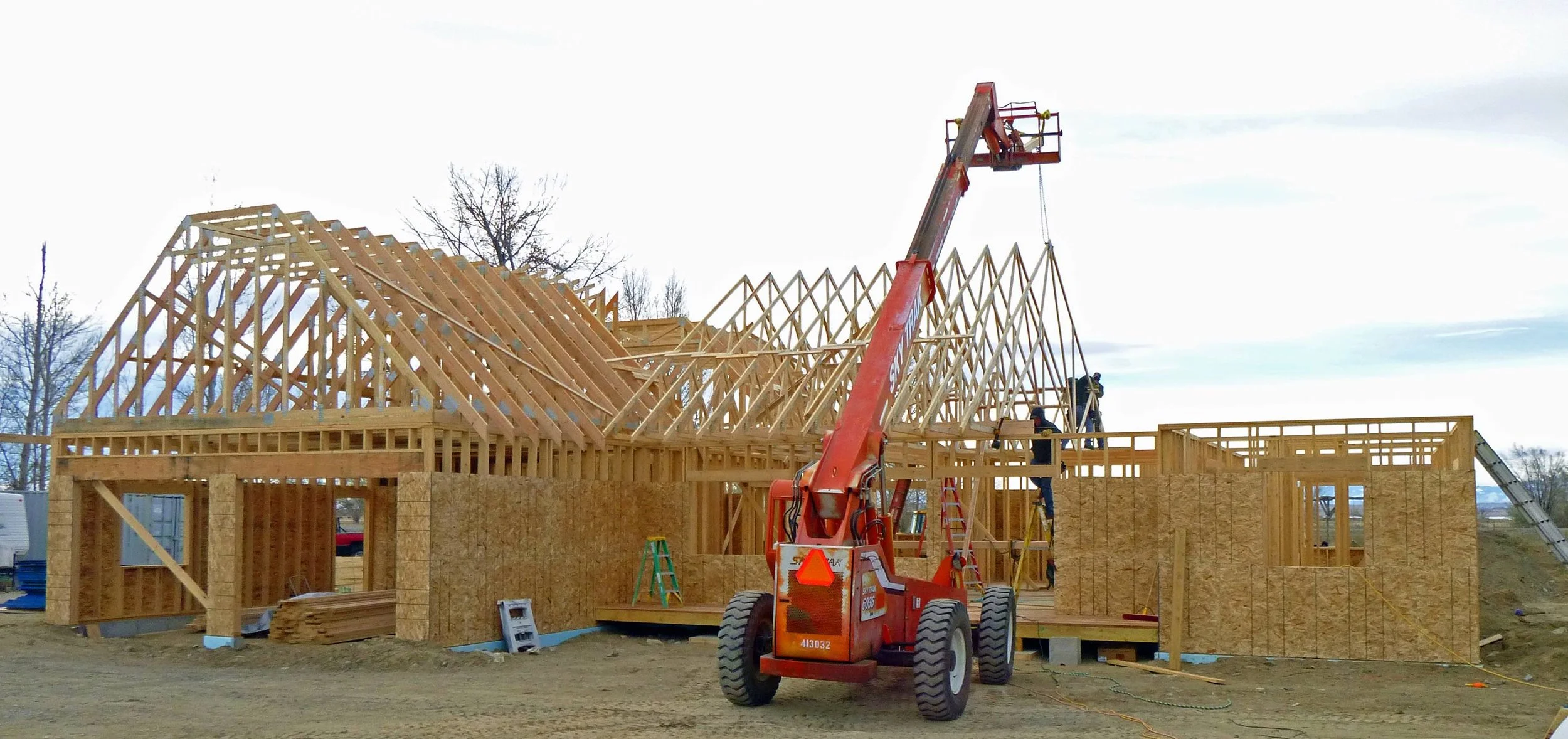 Construction site with a wooden house frame under construction, a red crane lifting materials, and workers installing the roof trusses.