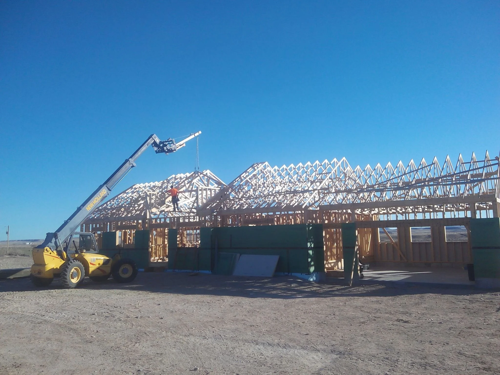 Construction site with a wooden house frame being built under a clear blue sky, a worker on site, and a crane lifting materials.