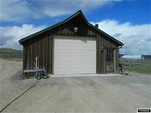 A small wooden garage with a white roll-up door, set on a dirt area with a cloudy sky in the background.