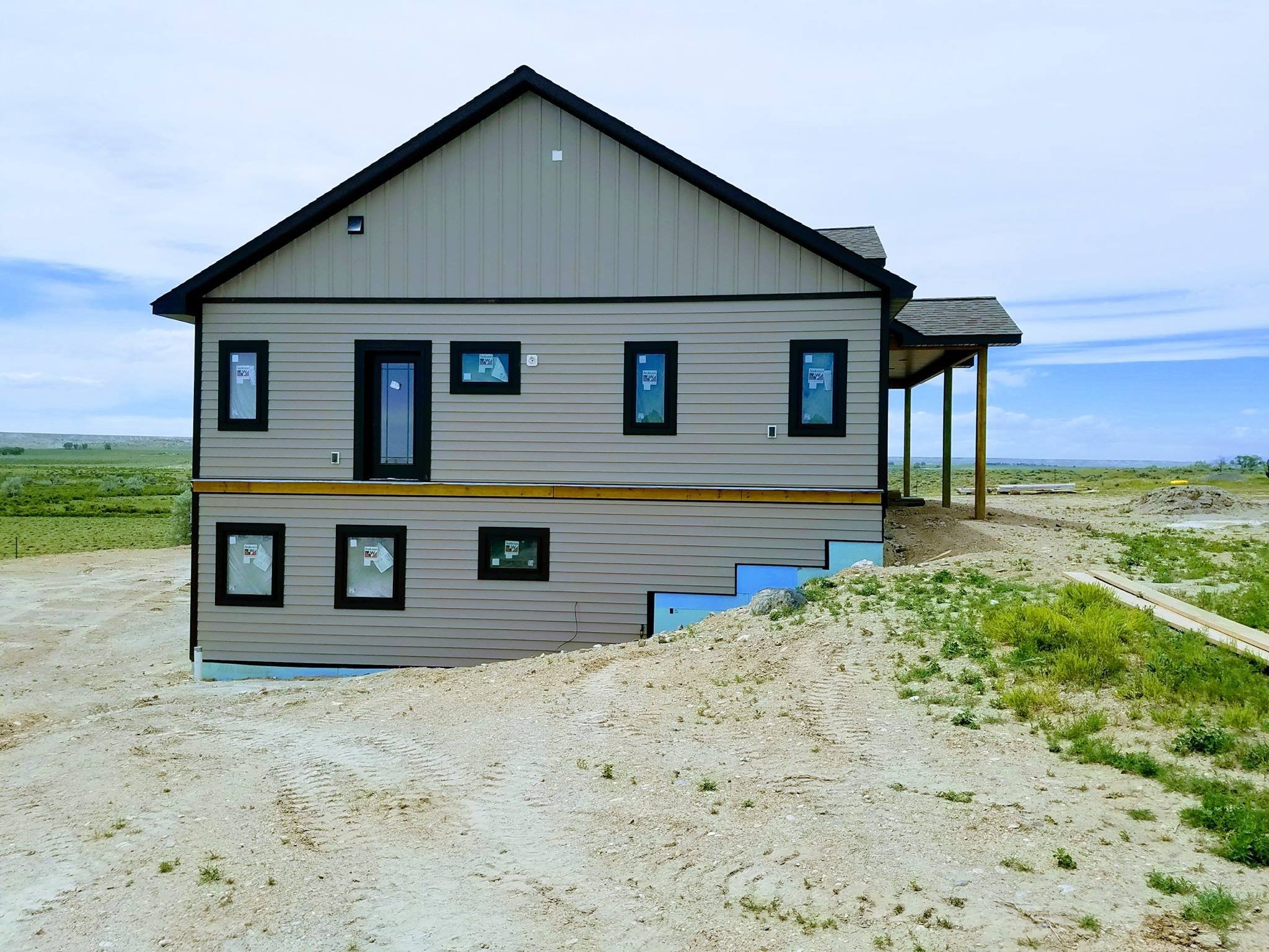 A two-story house under construction in a rural area, with beige and black siding, multiple windows, and a partially completed porch.