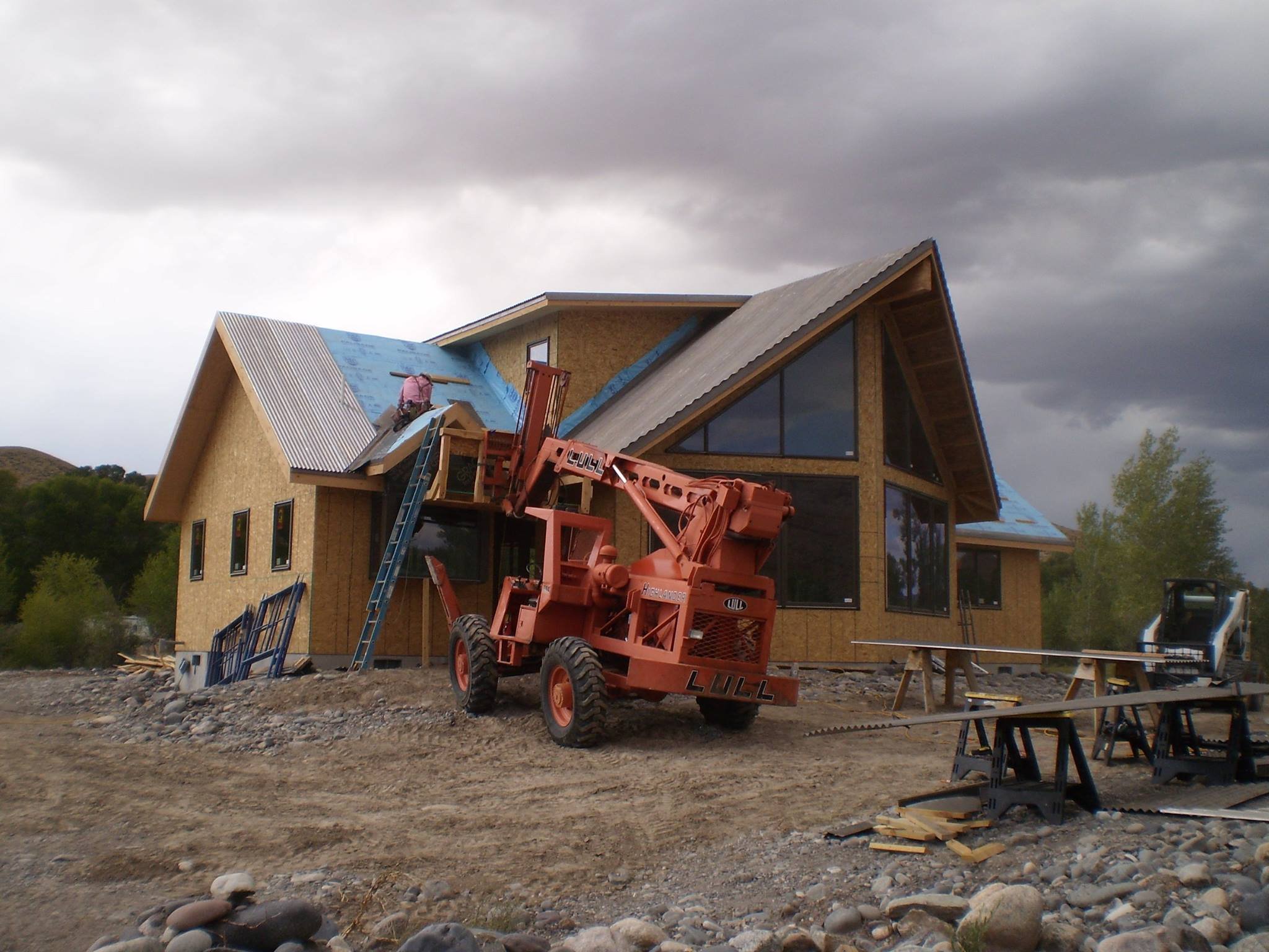 A house under construction with a worker installing roofing panels. Construction equipment and tools are in the foreground and there are cloudy skies above.