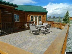 Outdoor patio with a dining table and chairs on a stone-paved surface, adjacent to a wooden house with a green roof.