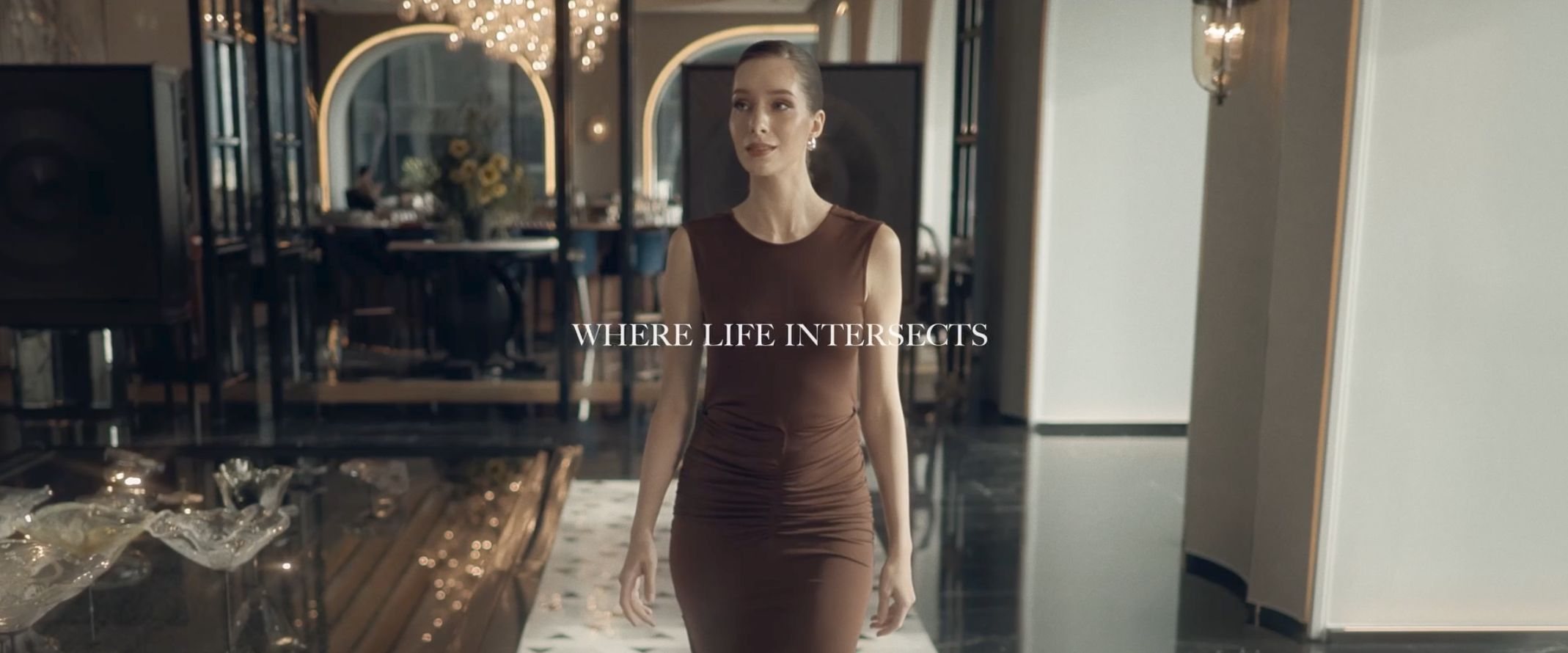 A woman in a fitted brown dress walking in an elegant room with large mirrors, chandelier, and floral arrangements, with the text 'Where Life Intersects' overlaid. One bangkok film. The Ritz Carlton bangkok film.