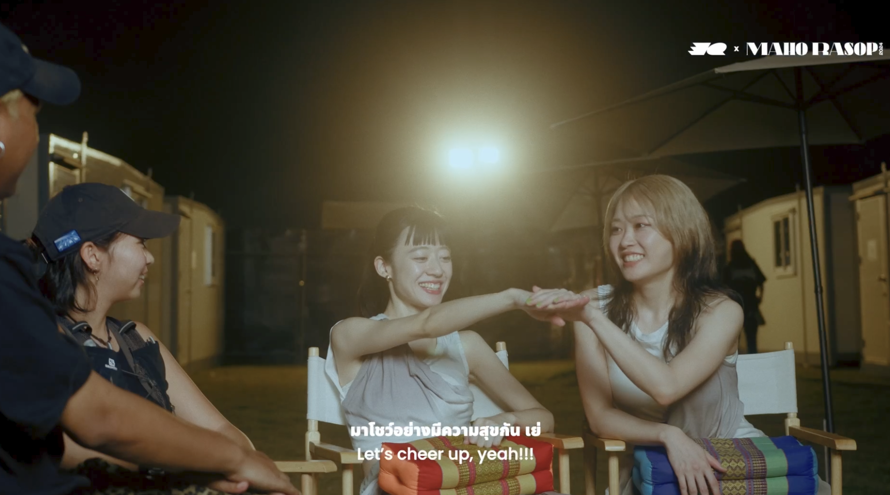 Three women sitting outdoors at night, smiling and engaging in a cheer gesture, with a man nearby and portable toilets in the background, under umbrellas. Music event and festival video. Interview bands