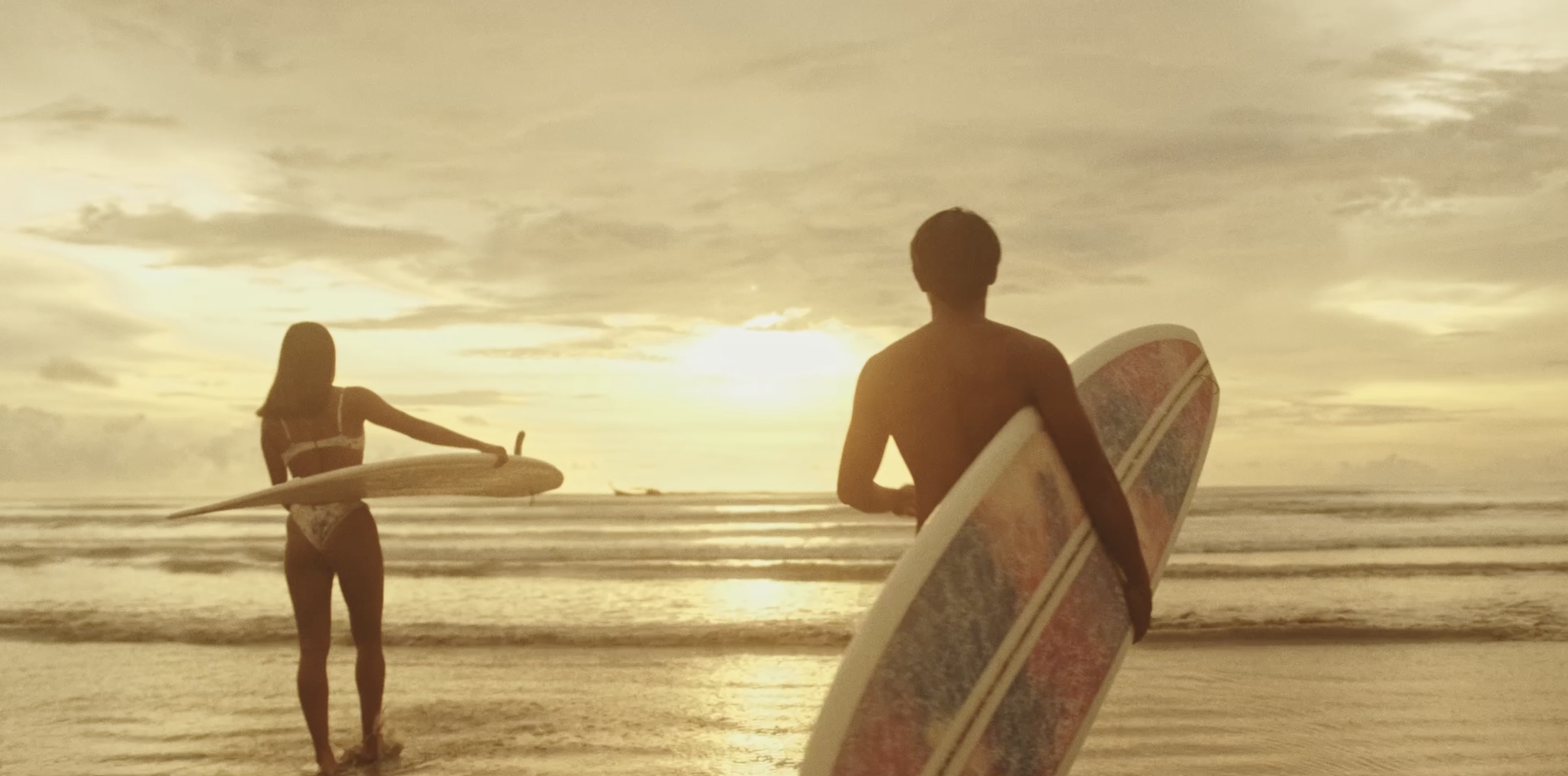 Silhouettes of two surfers walking on the beach with surfboards at sunset, ocean waves in the background, clouds in the sky, and golden lighting.