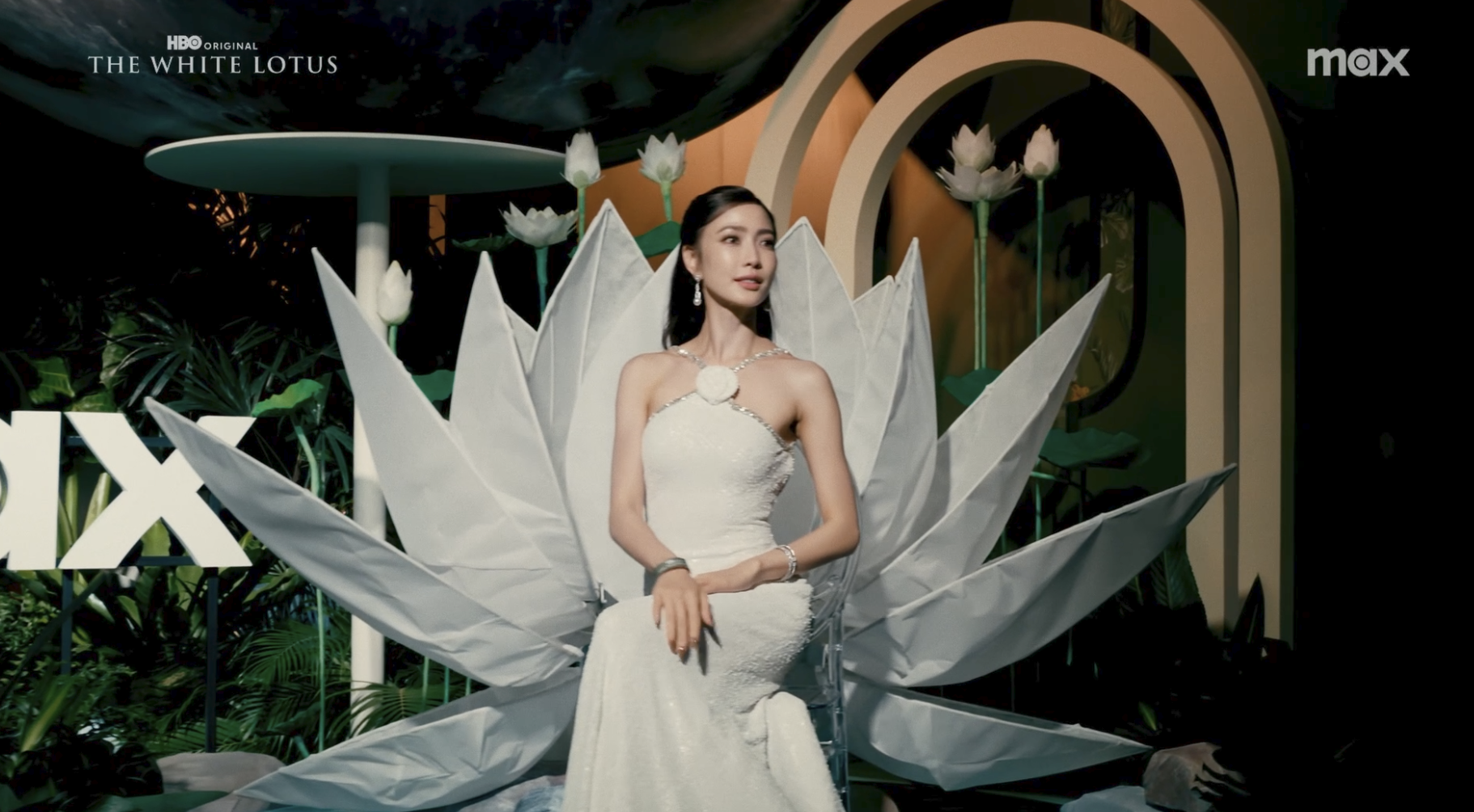 A woman in a white dress sitting on a clear chair in front of a large lotus flower display, with a green and black background, and a sign that reads 'X' on the left, in an ornate setting.
Film opening, promotional video. The white lotus event.