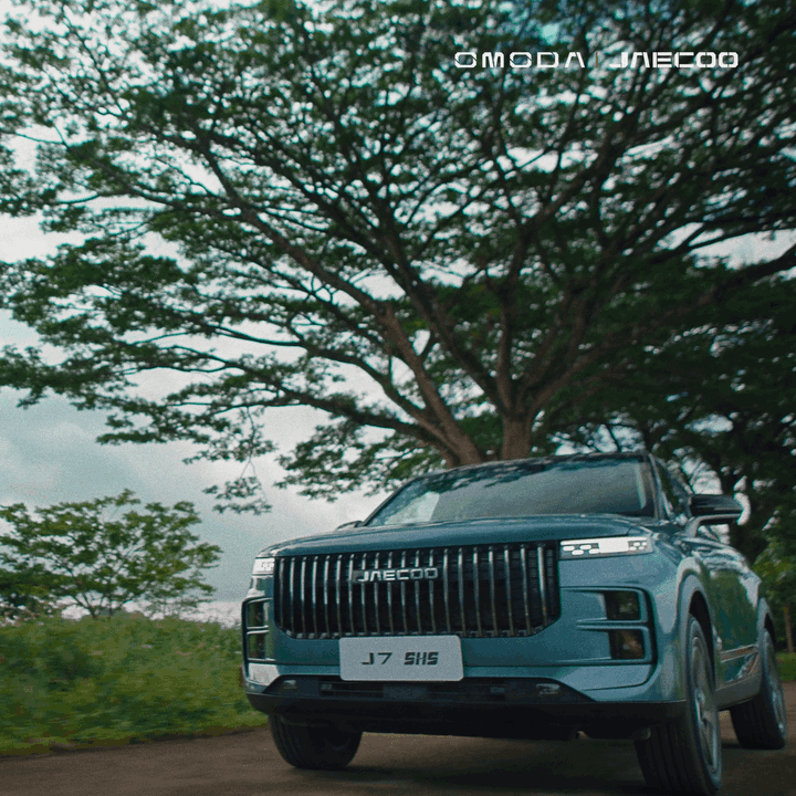 Blue JAECOO J7 SHS SUV parked on a dirt road with a large leafy tree and green foliage in the background.