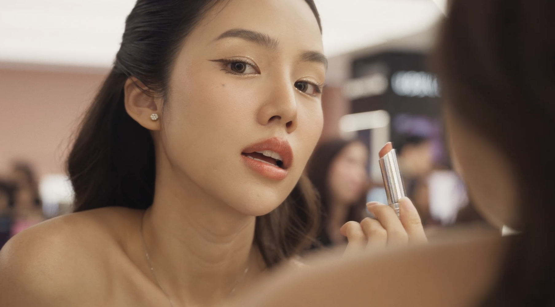 A woman applying lipstick in a makeup store, with her face turned toward a mirror, surrounded by other shoppers. High fashion brand. Make up and event. Central chitlom