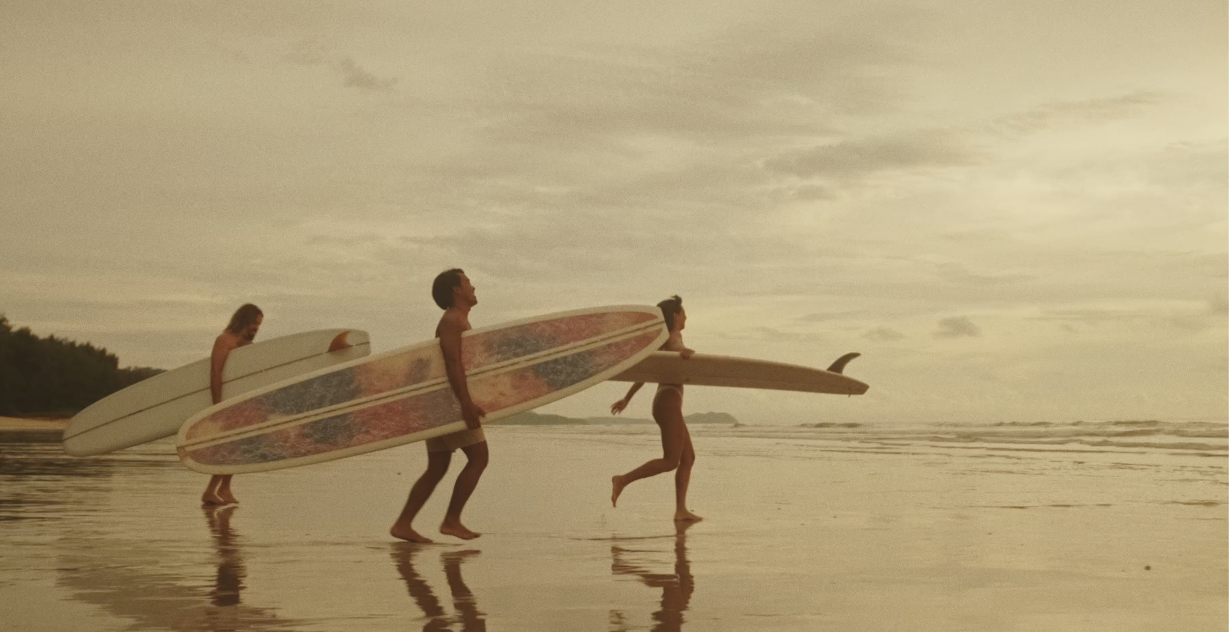 Three people walking on the beach with surfboards under sunset sky. Surf film, outdoor sunset indie film