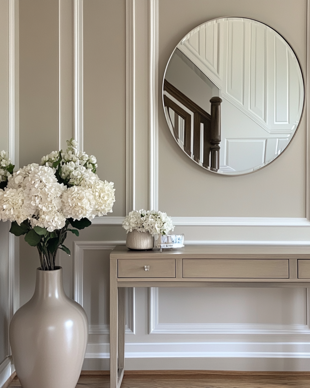 An indoor console table with two planters of white flowers in front of a wall with decorative molding and a round mirror reflecting part of a staircase and white paneled door.