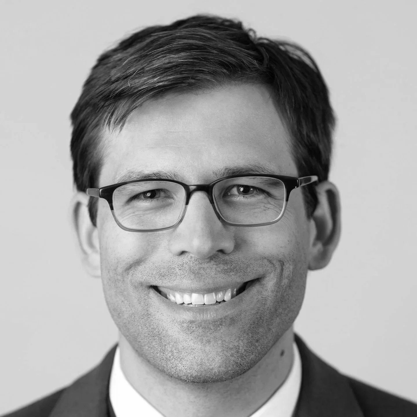 Black and white close-up portrait of a smiling man wearing glasses, dressed in a suit and tie, with neatly combed hair.