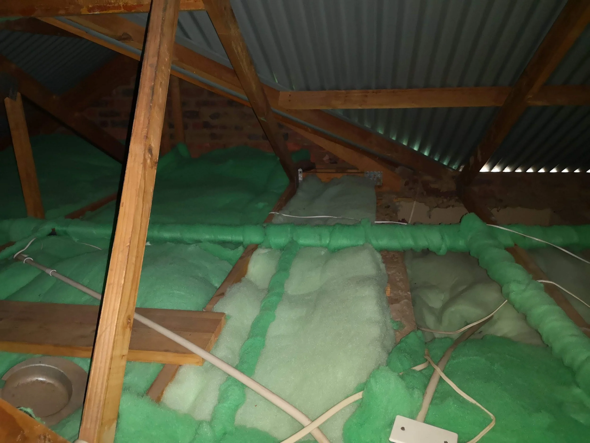 Insulation foam installation in an attic with wooden beams and a corrugated metal roof.