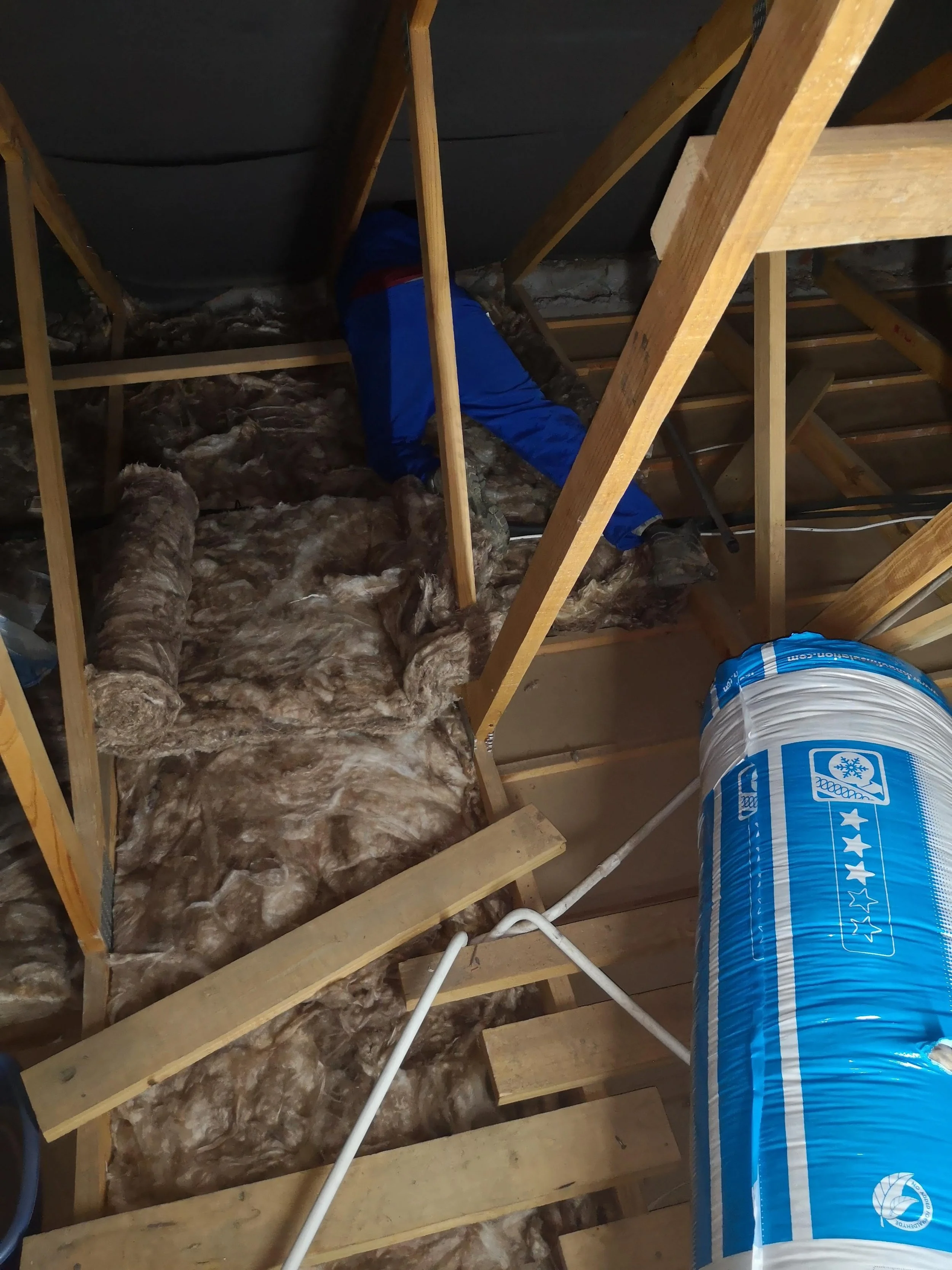 A person in blue and red clothing working on insulation in the attic, with exposed wooden framing and ductwork.