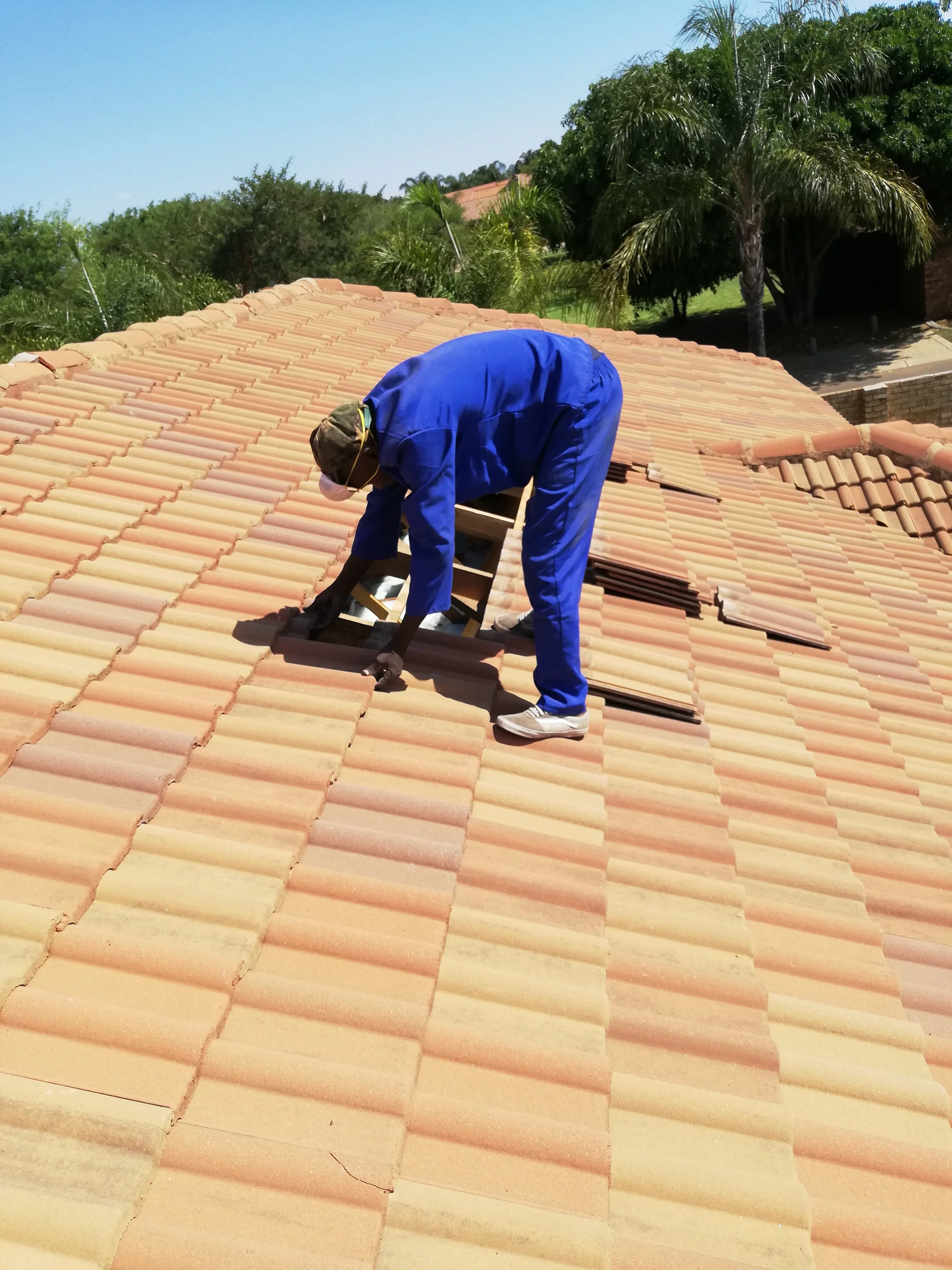 A worker in blue overalls and a cap is repairing a tiled roof under a clear sky, surrounded by green trees.