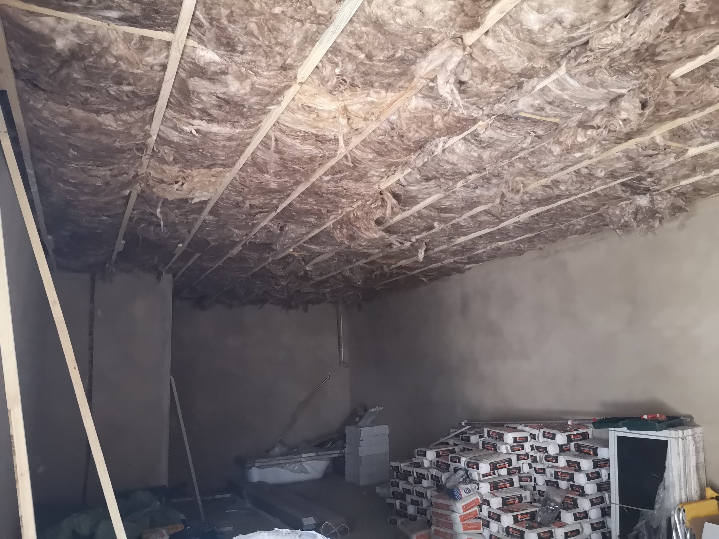 Interior room under construction with exposed insulation on the ceiling, cinder blocks stacked against the wall, and construction tools and materials scattered around.