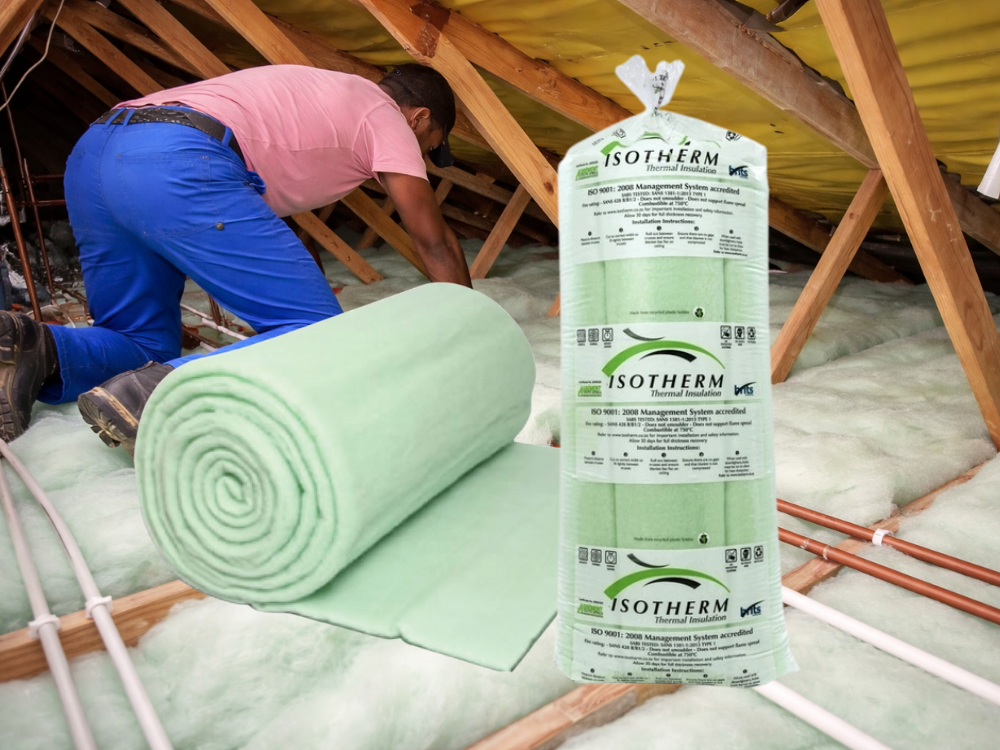 A person installing green thermal insulation rolls in an attic with exposed wooden beams and pipes.