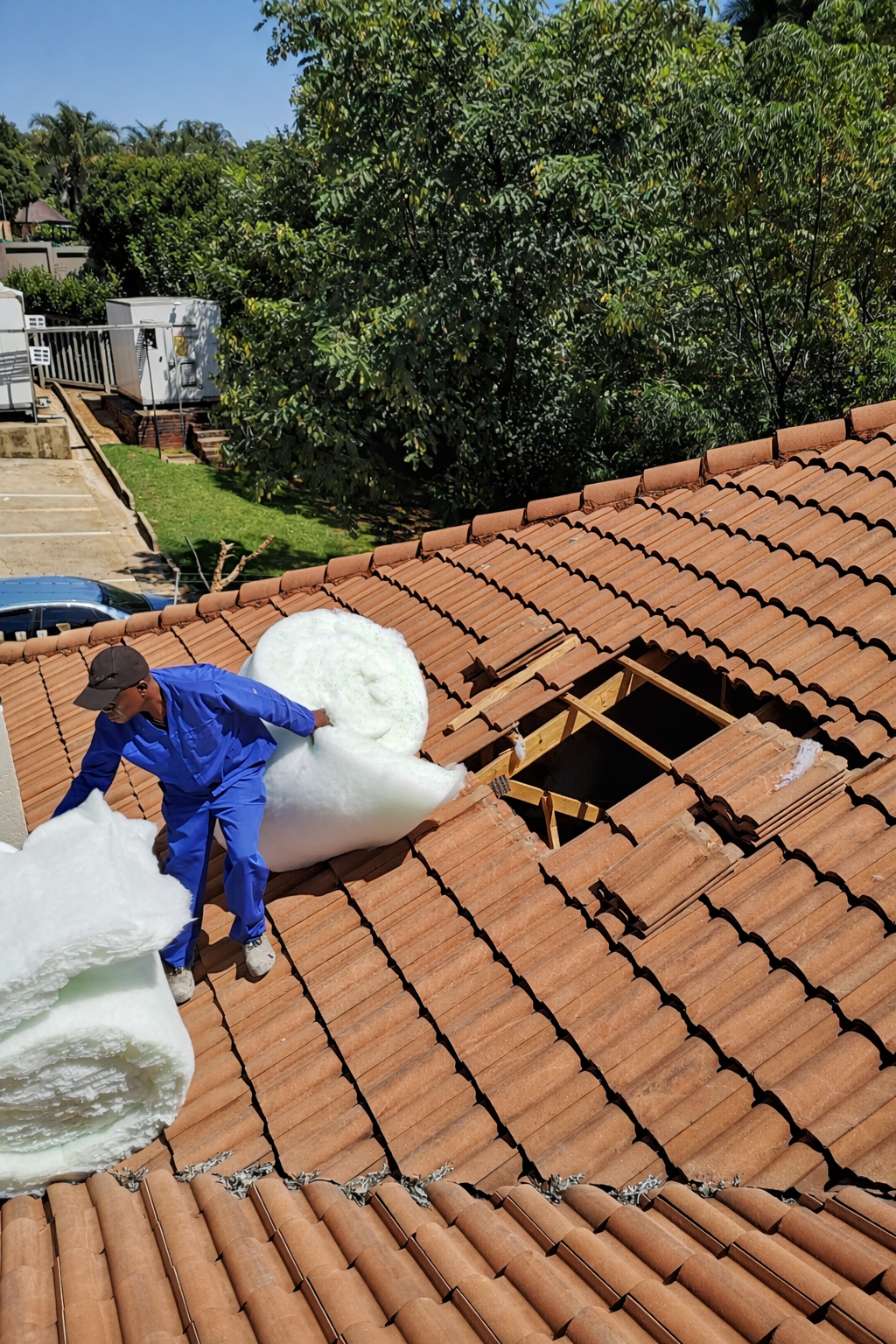 A worker in blue overalls and a cap installing insulation foam on a tiled roof with a large section cut out.