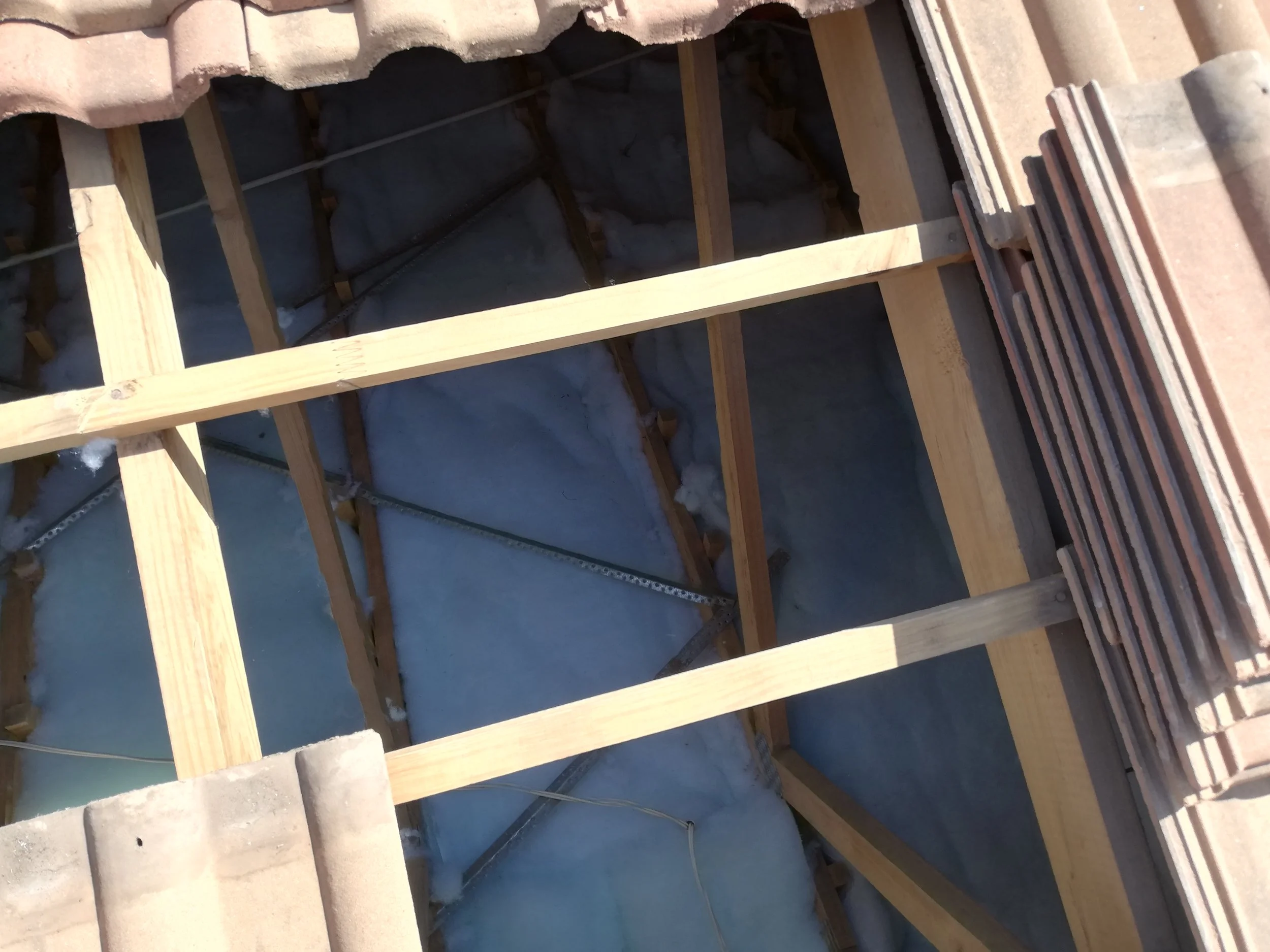 View of a roof during construction showing wooden framing, roofing tiles stacked on the edge, and some insulation material visible inside the open roof area.