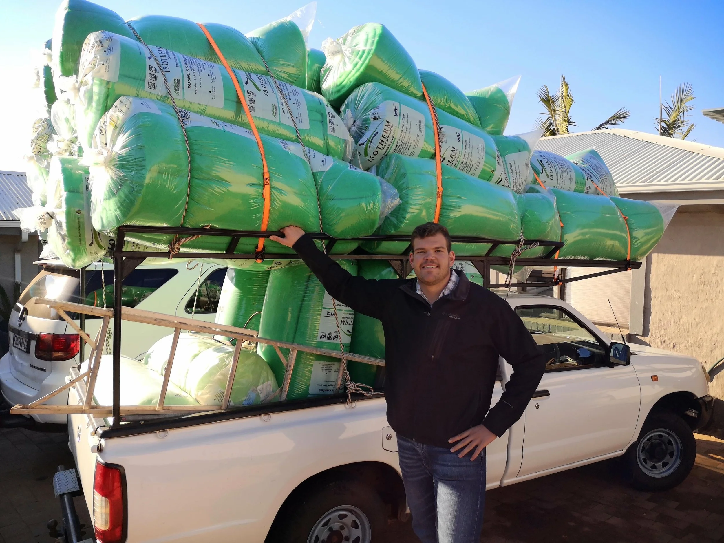 A man standing next to a white pickup truck loaded with large green bags of potting soil, secured with orange straps. The man is wearing a black jacket and jeans, smiling at the camera, with a suburban house and palm trees in the background under a c