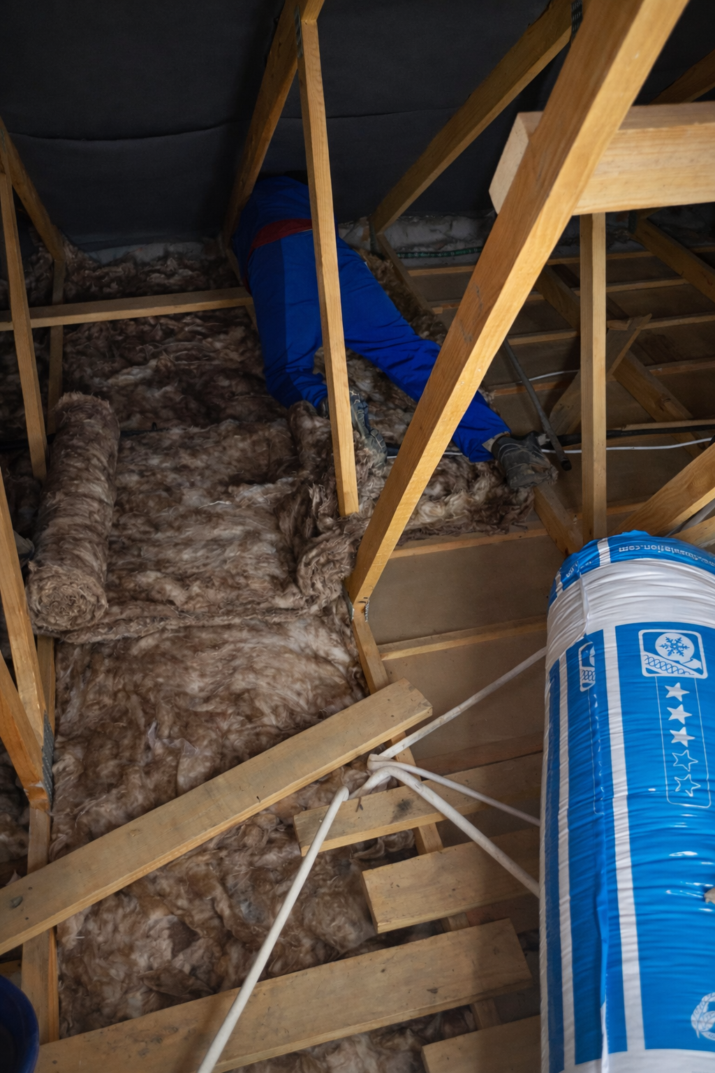 A person working on insulation in an attic, surrounded by wooden beams and insulation material, with a large roll of insulation nearby.