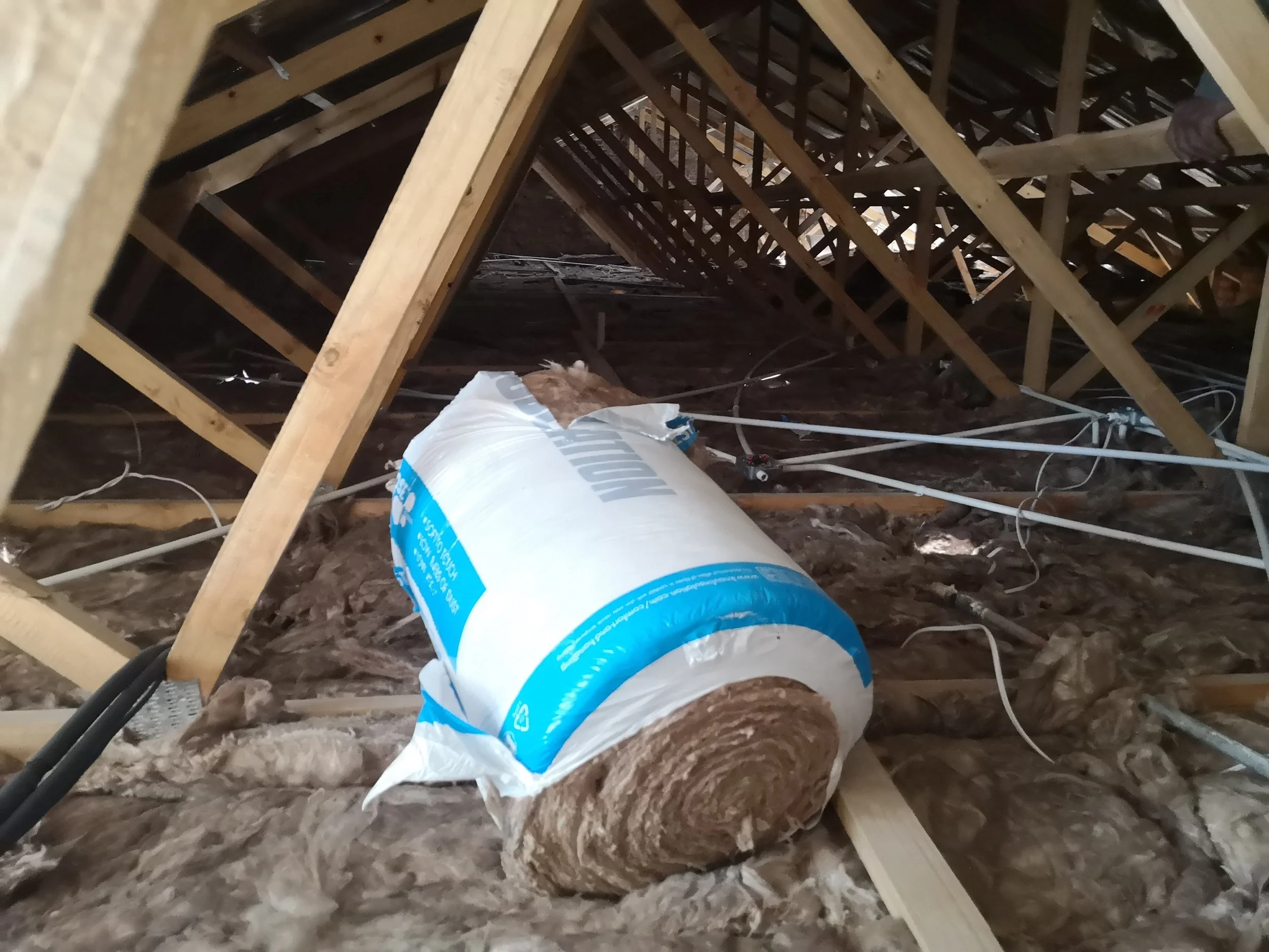 Insulation roll on attic floor with exposed wooden roof trusses and electrical wiring.