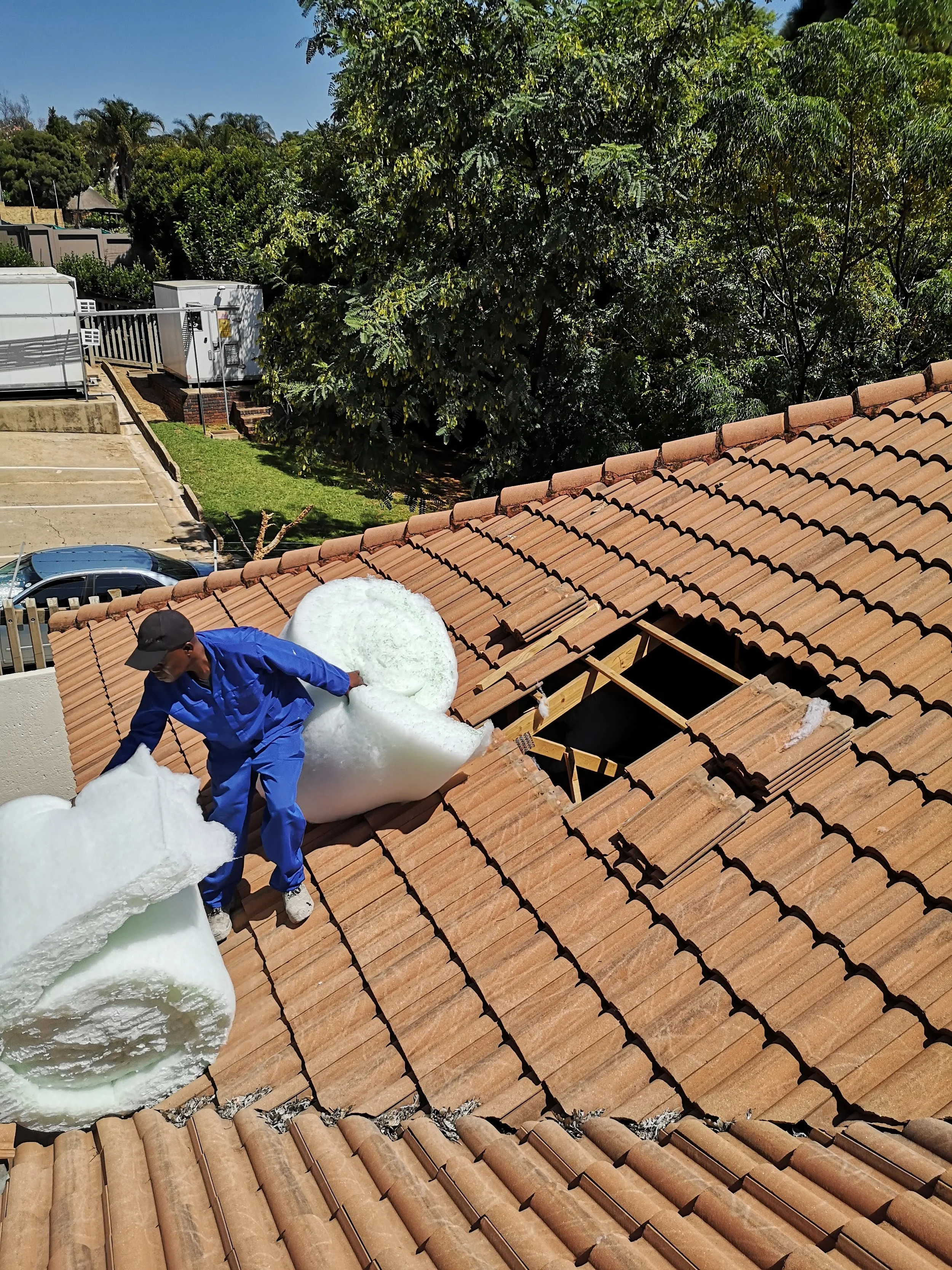 A person in blue work overalls and a black cap placing a large piece of white insulation foam on a tiled roof with a hole. There are trees and parked cars in the background.