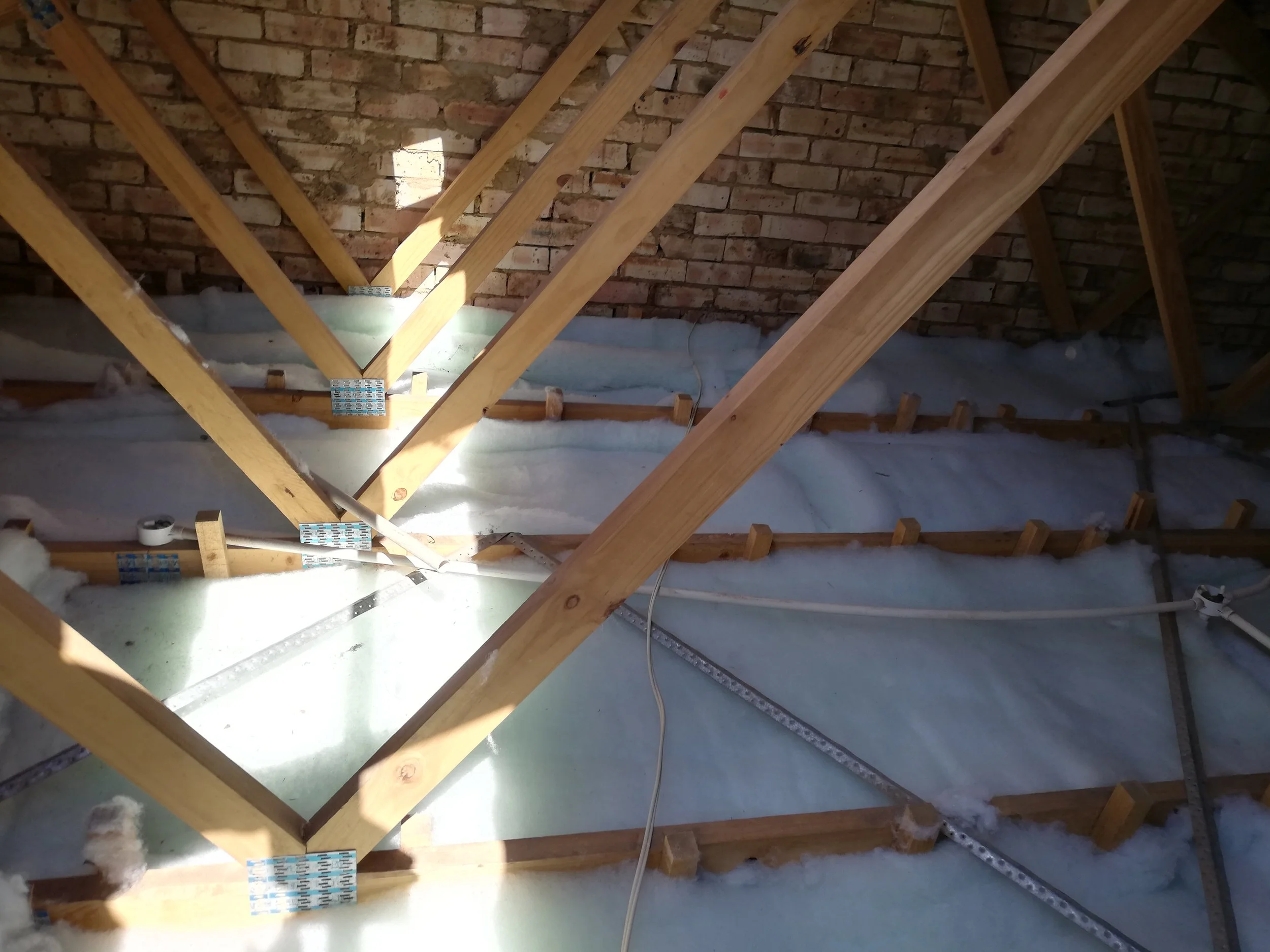 Construction of attic with wooden beams, insulation, and brick wall in the background.
