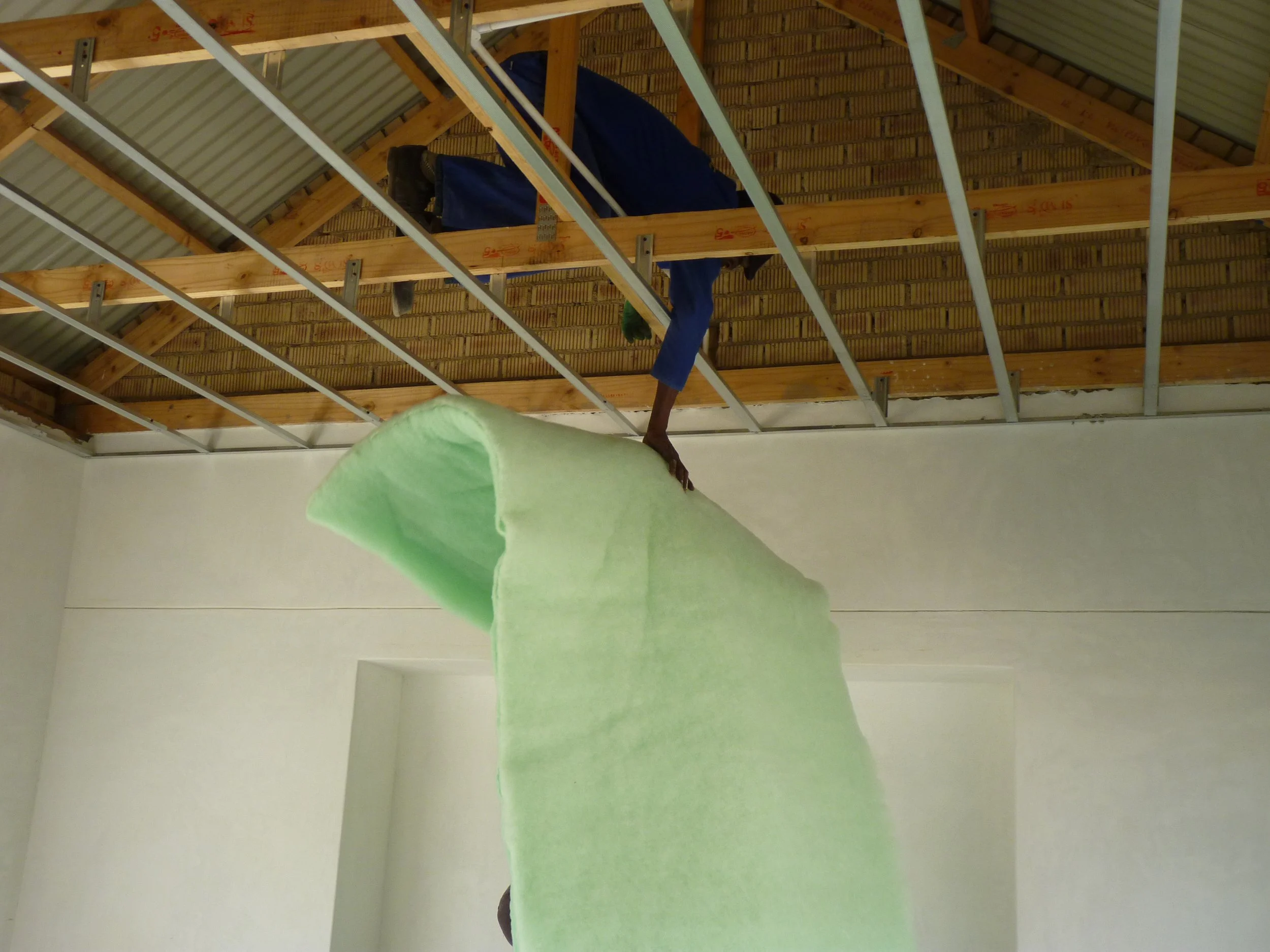 Person working on constructing a ceiling, hanging upside down from the ceiling beams, with a green insulation roll.