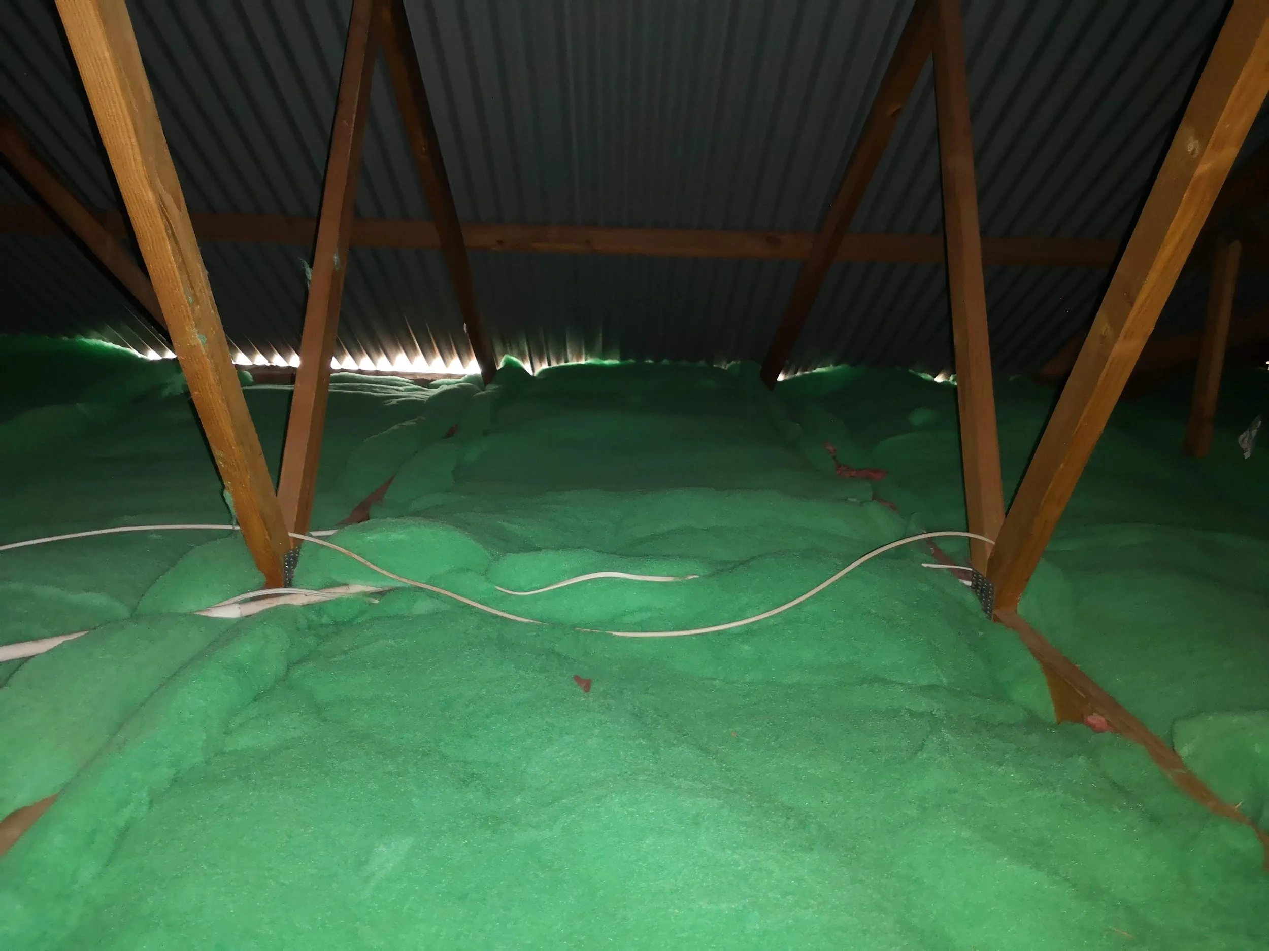 Attic space with green insulation foam and wooden beams, with some electrical wiring visible.