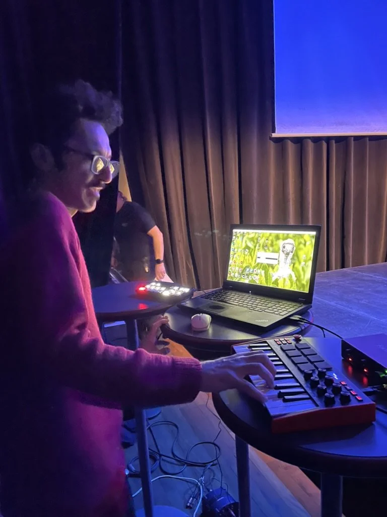Person playing a small electronic keyboard with a laptop and other electronic music equipment on a table in a dark room with stage curtains.