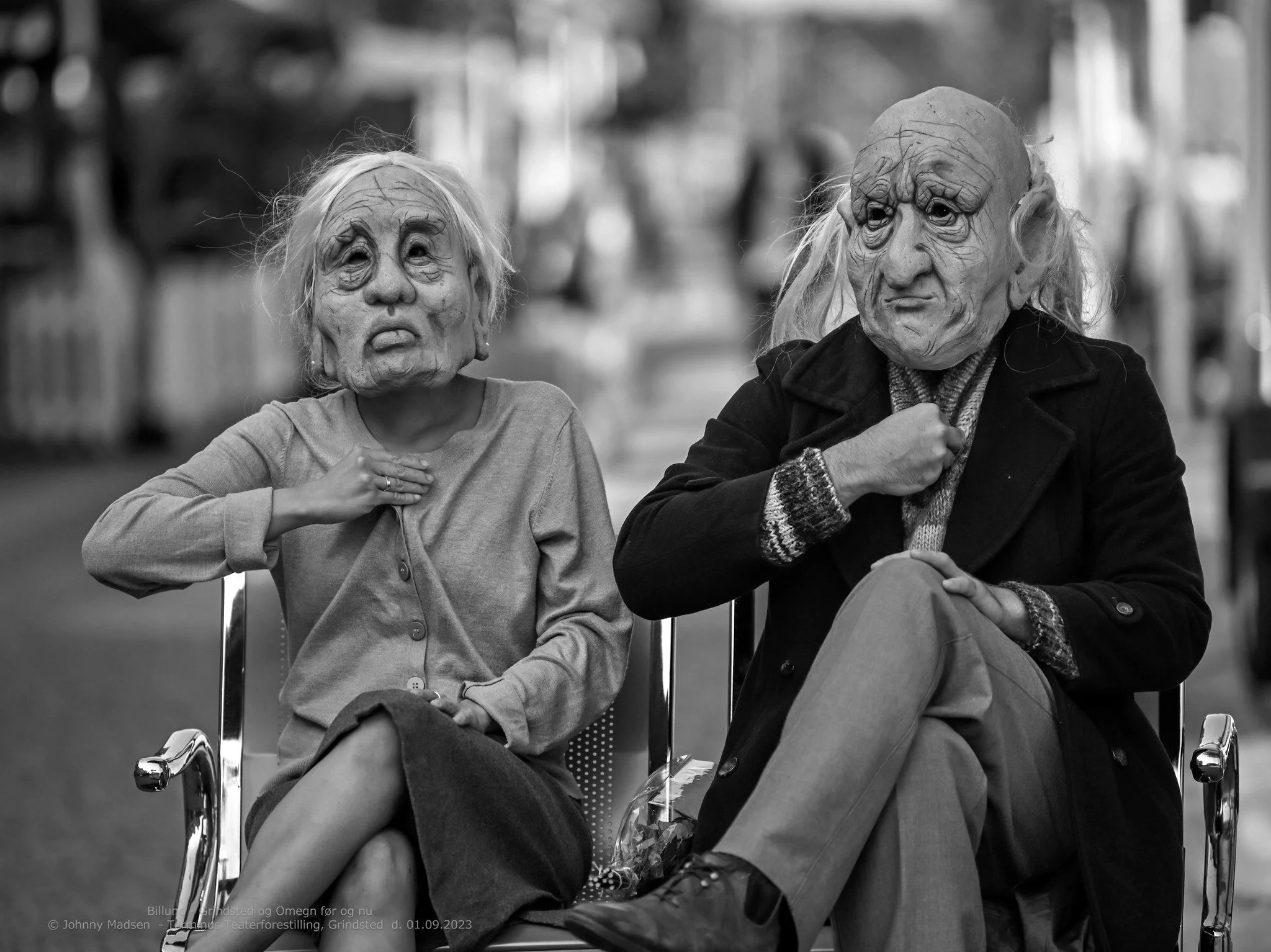 Black and white photo of two people wearing grotesque masks resembling elderly individuals, sitting in chairs outdoors. The person on the left is wearing a light-colored cardigan, with one hand on their chest, the other on their lap, and crossed legs