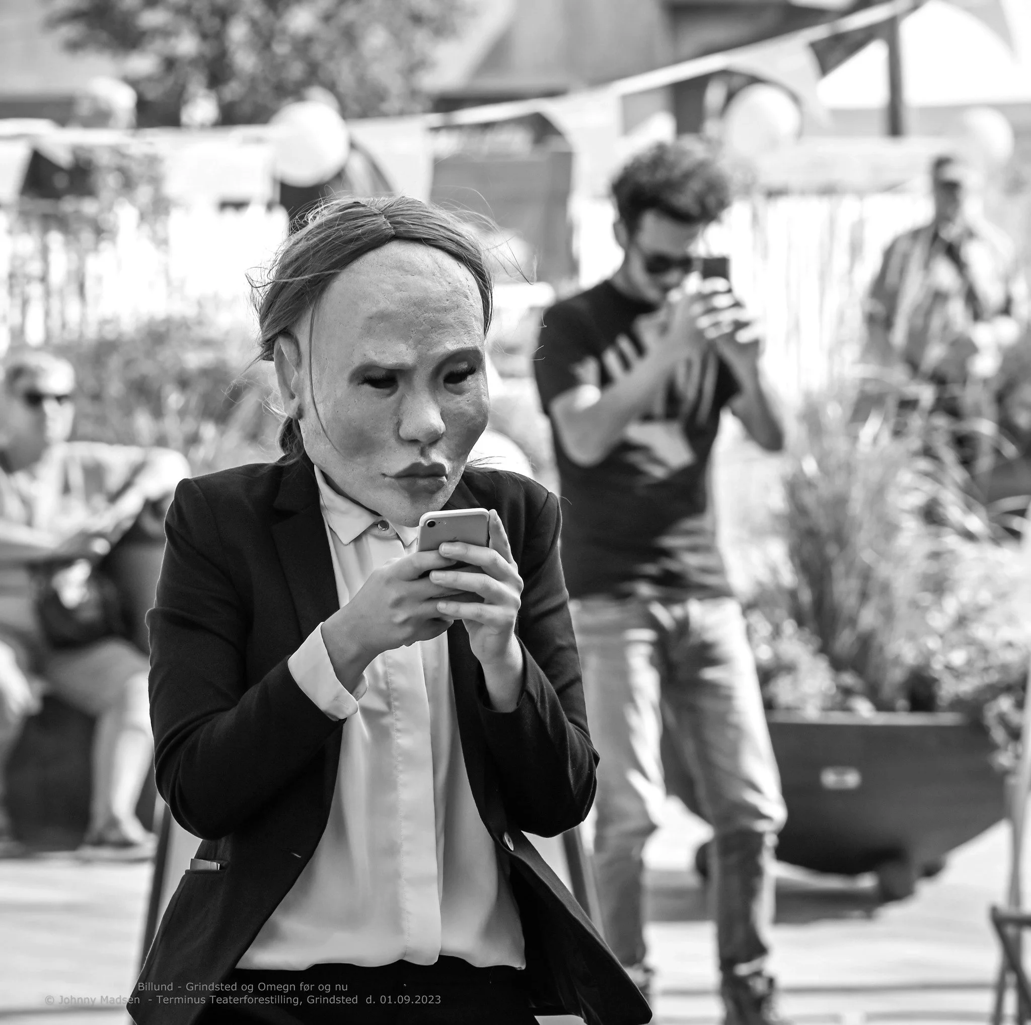 A black and white photo of a woman in a blazer and white shirt looking at her phone, with people in the background also using their phones, outdoors during daytime.