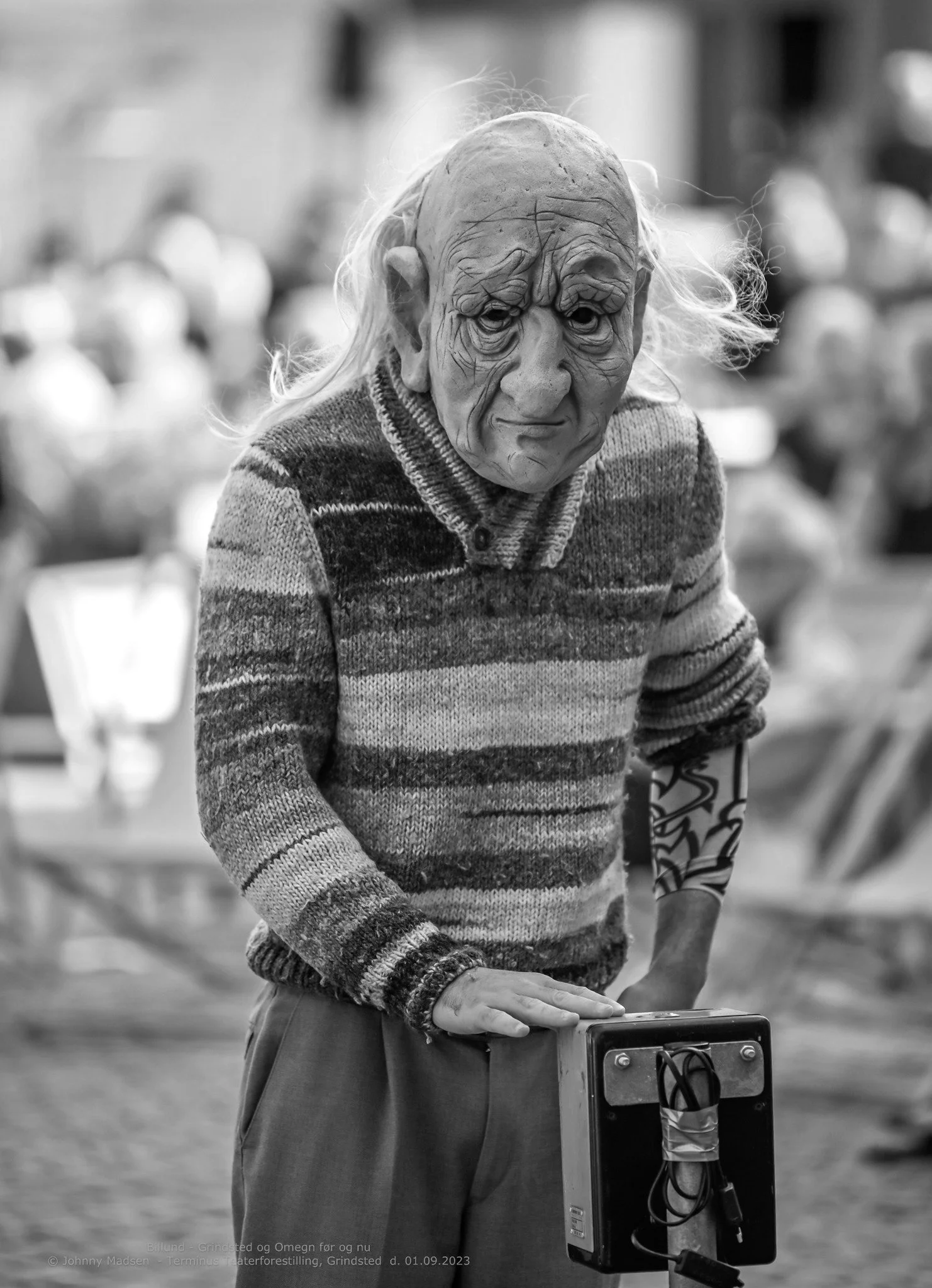 A person wearing a realistic mask resembling an old, wrinkled face with long hair, dressed in a striped sweater, standing outdoors and leaning on a small case or box.