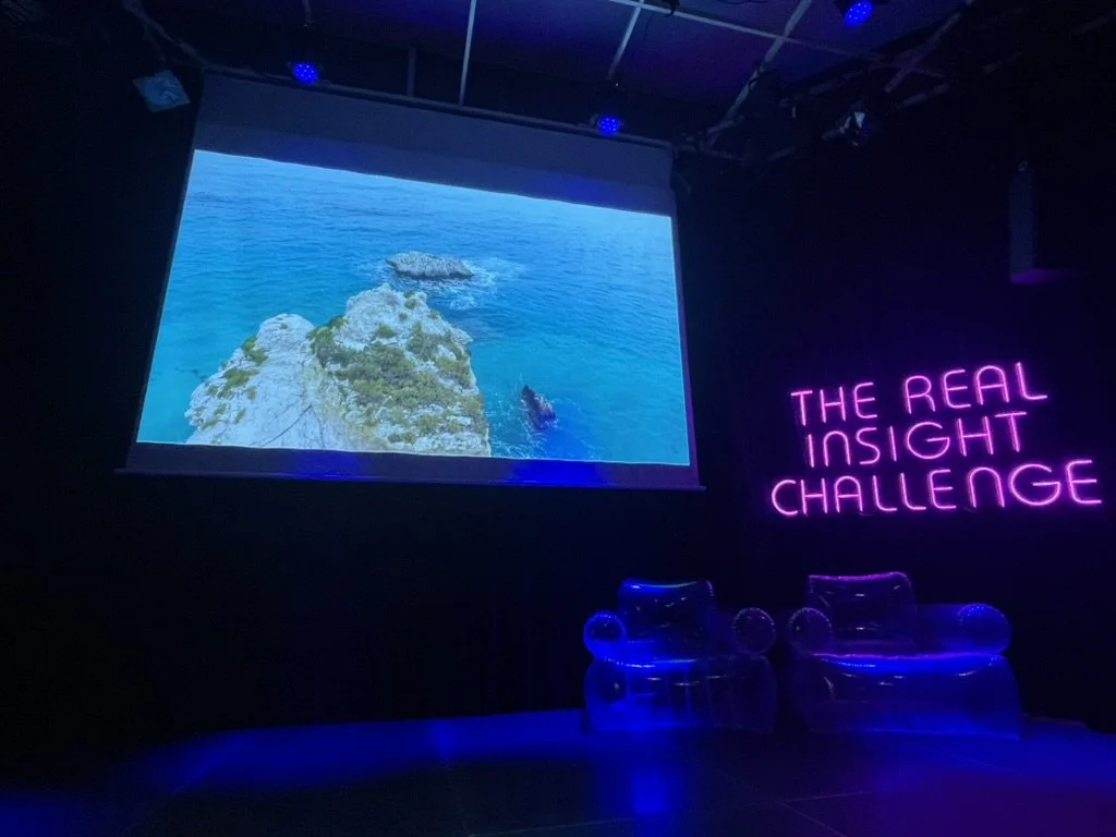 Neon pink sign reading 'The Real Insight Challenge' next to an inflatable transparent chair, with a large screen displaying a seascape with rocks and an ocean scene.