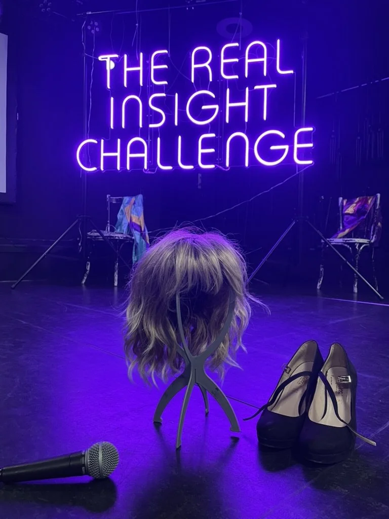 Stage with neon purple sign reading 'The Real Insight Challenge', a wig on a stand, high heel shoes, and a microphone on the floor.