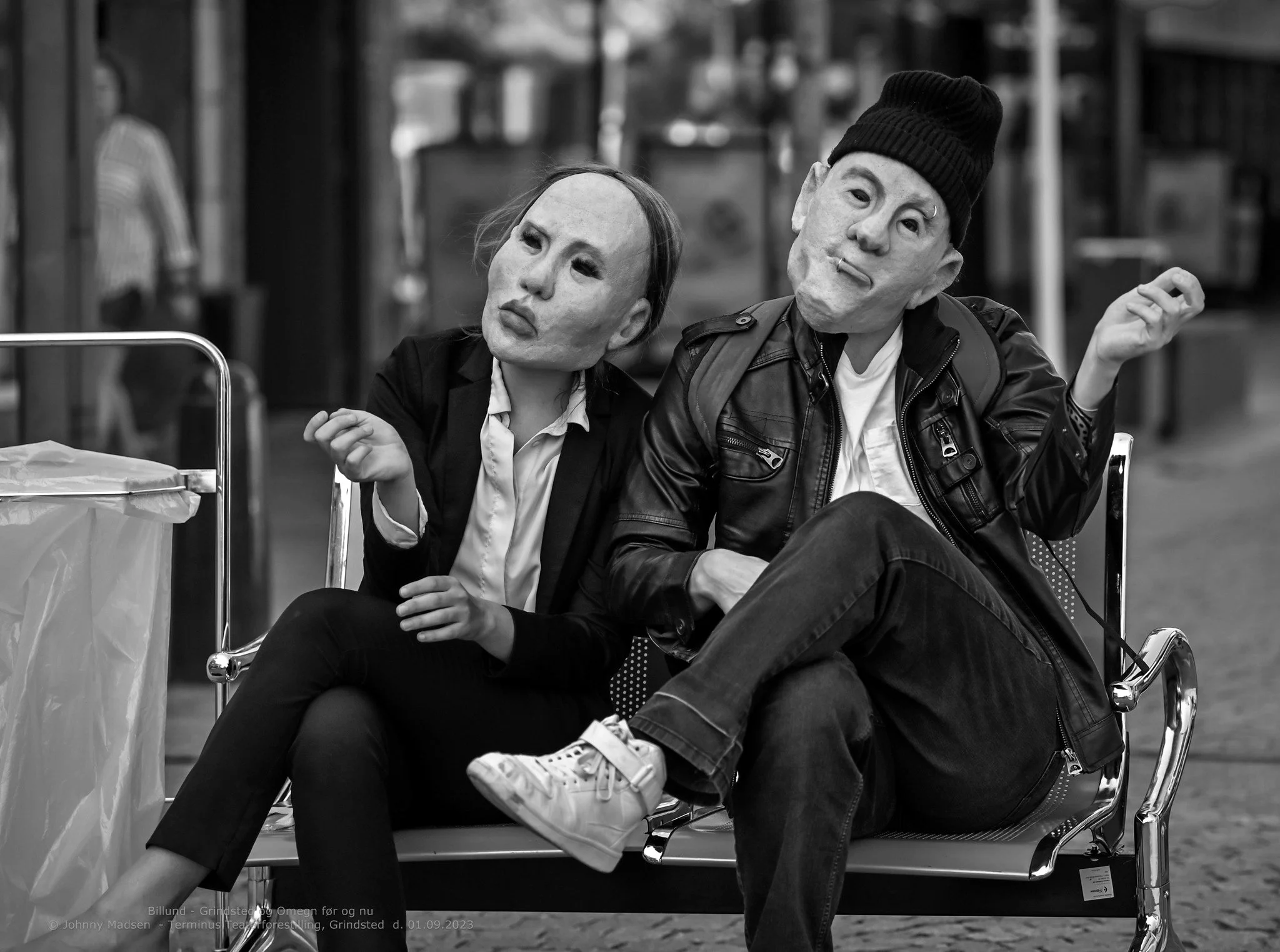 Black and white photo of two people sitting on a bench with masks that resemble exaggerated, artistic faces. The person on the left is wearing formal attire, and the person on the right is dressed casually in a leather jacket and beanie, with a cigar