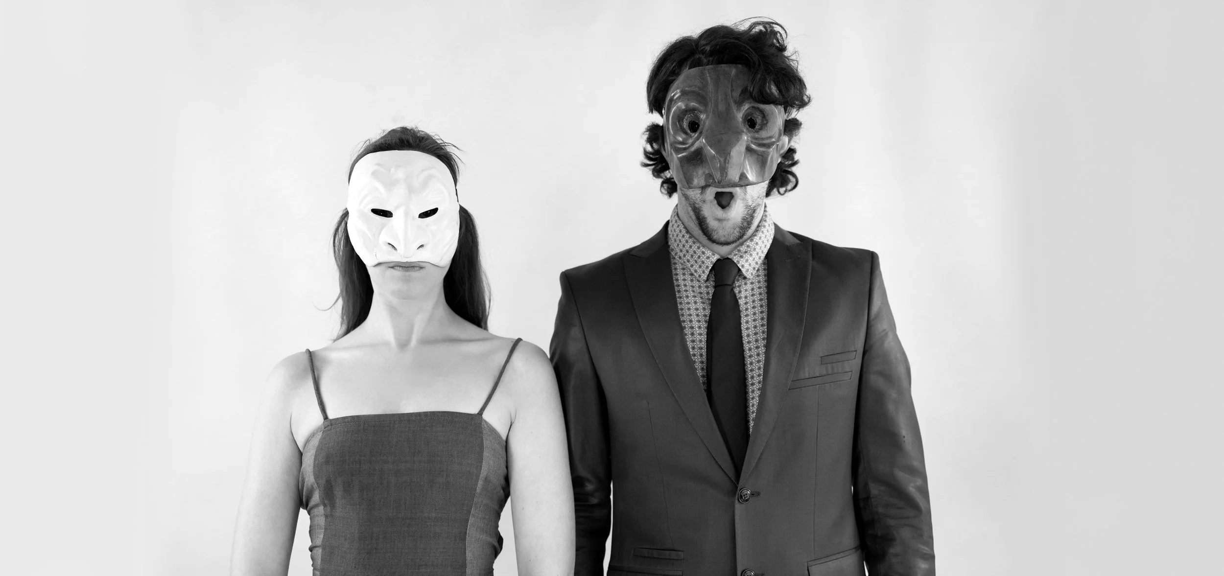 Man and woman wearing masks in front of plain wall, man in suit and woman in dress
