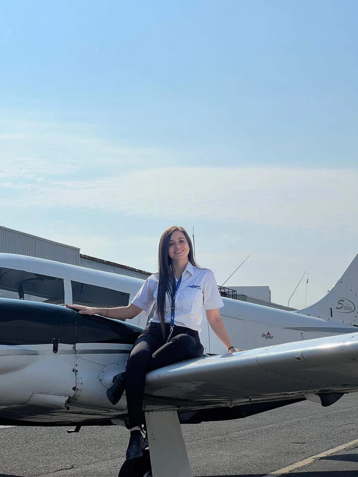Mujer sentada sobre un avión ligero en una pista de aeropuerto, con vestido blanco y pantalones negros, sonriendo, en un día despejado.
