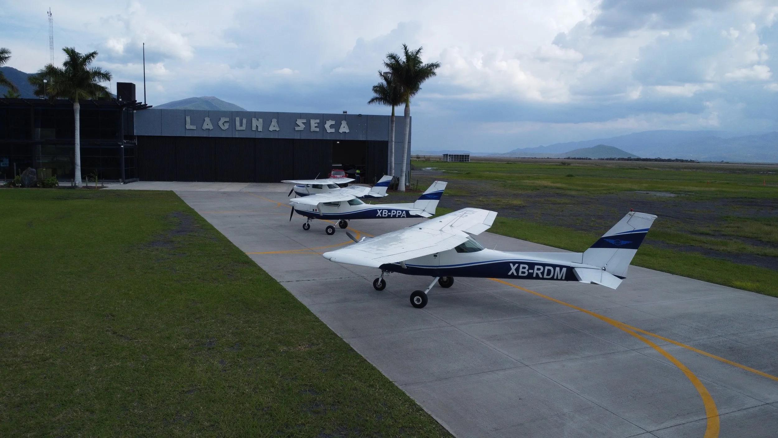 Tres avionetas pequeñas en la pista del aeropuerto junto a un edificio que dice 'Laguna Seca', con montañas y cielos nublados en el fondo.
