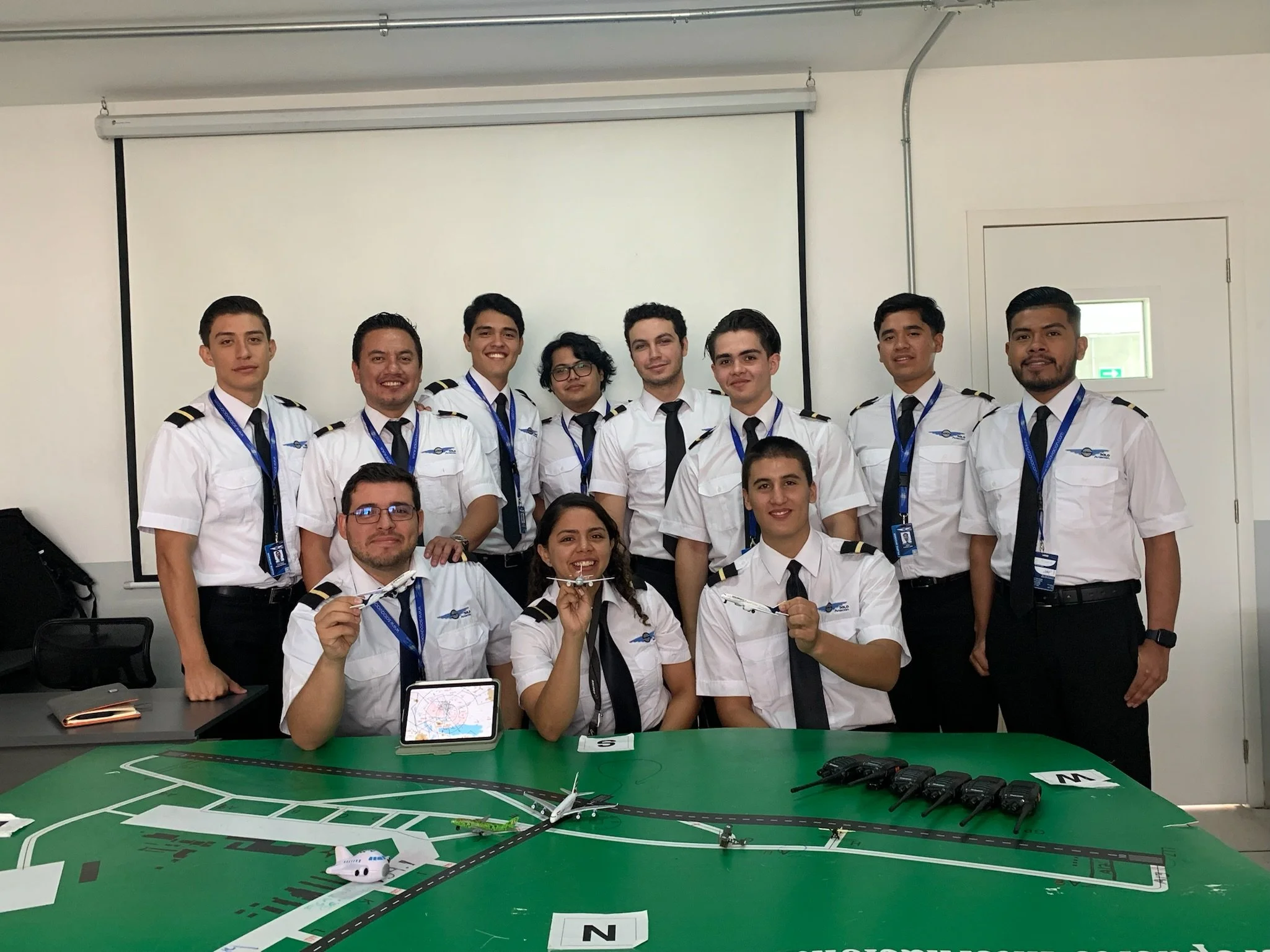 Grupo de personas vestidas con uniforme de pilotos, posando en una sala de clases con una maqueta de aeropuerto y aviones.