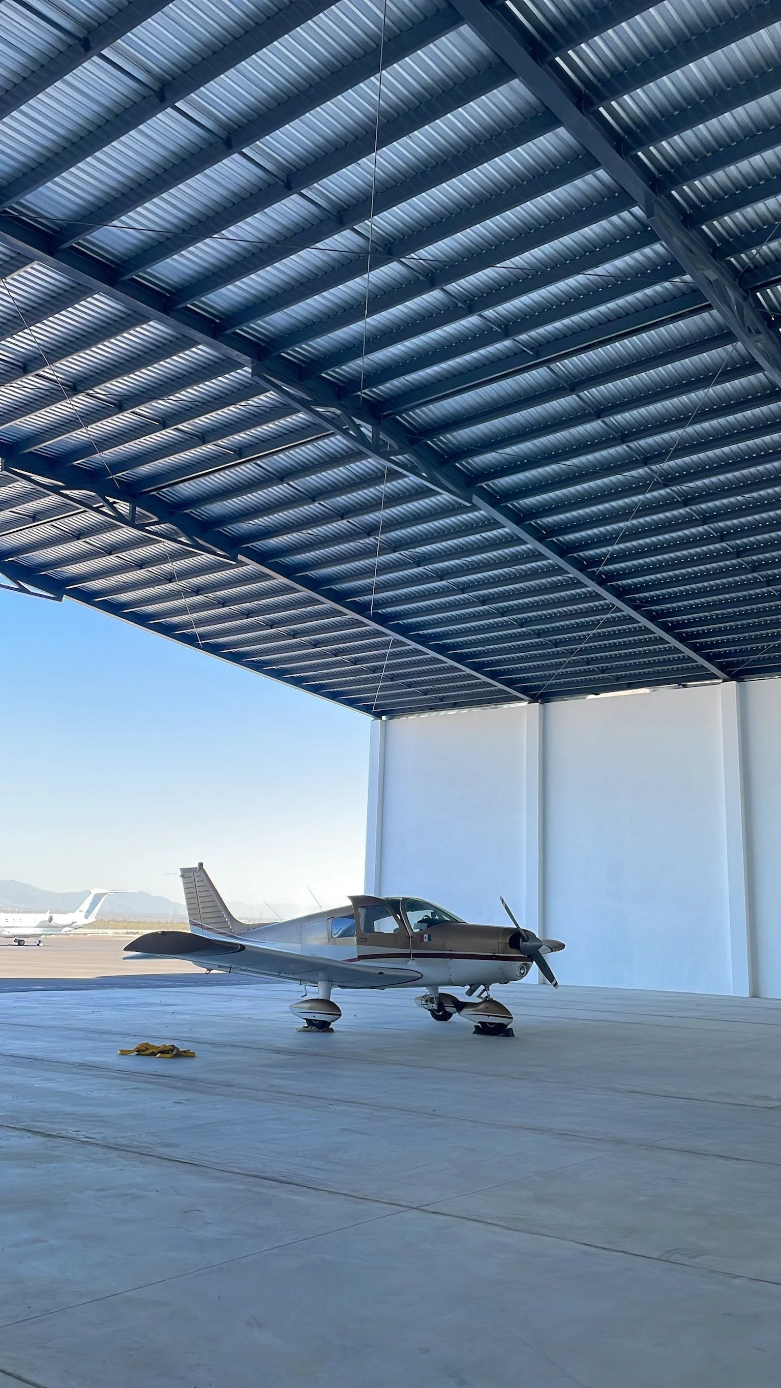 Avión pequeño en un hangar con estructura de metal y techo de láminas, vista hacia la pista del aeropuerto en el fondo.