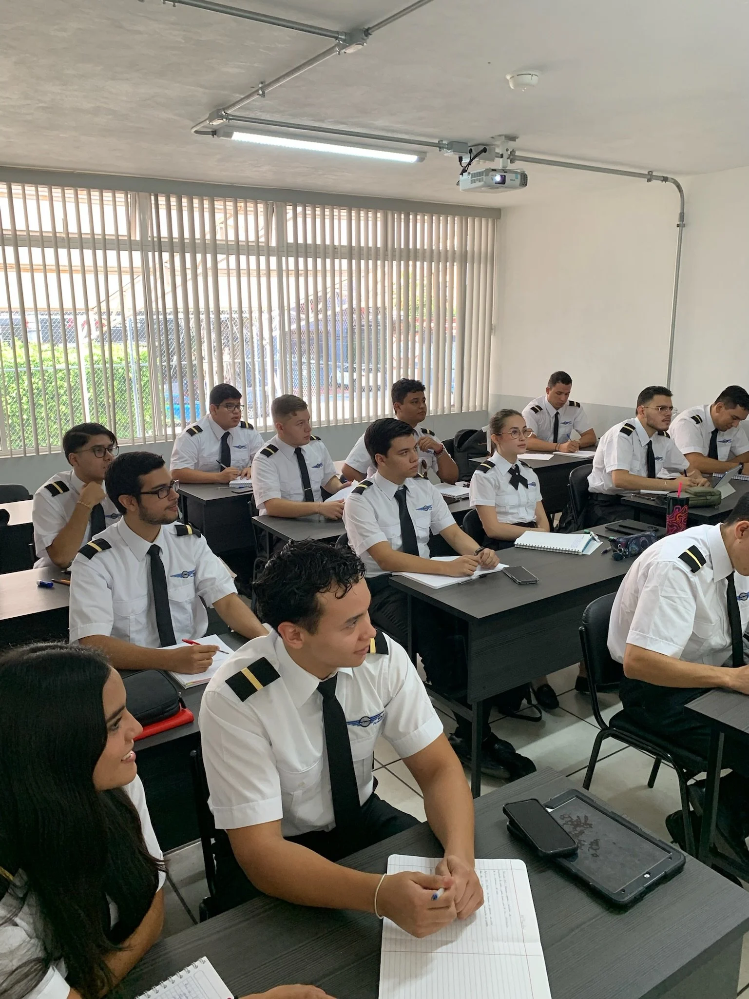 Estudiantes en uniforme de piloto en una clase o seminario en un aula con ventanas grandes y persianas verticales.