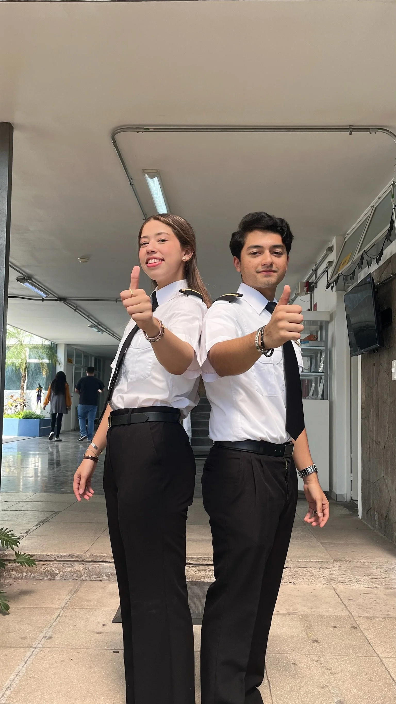 Dos jóvenes en uniformes de aerolínea posando y haciendo gestos de aprobación en un paso cubierto en un aeropuerto.