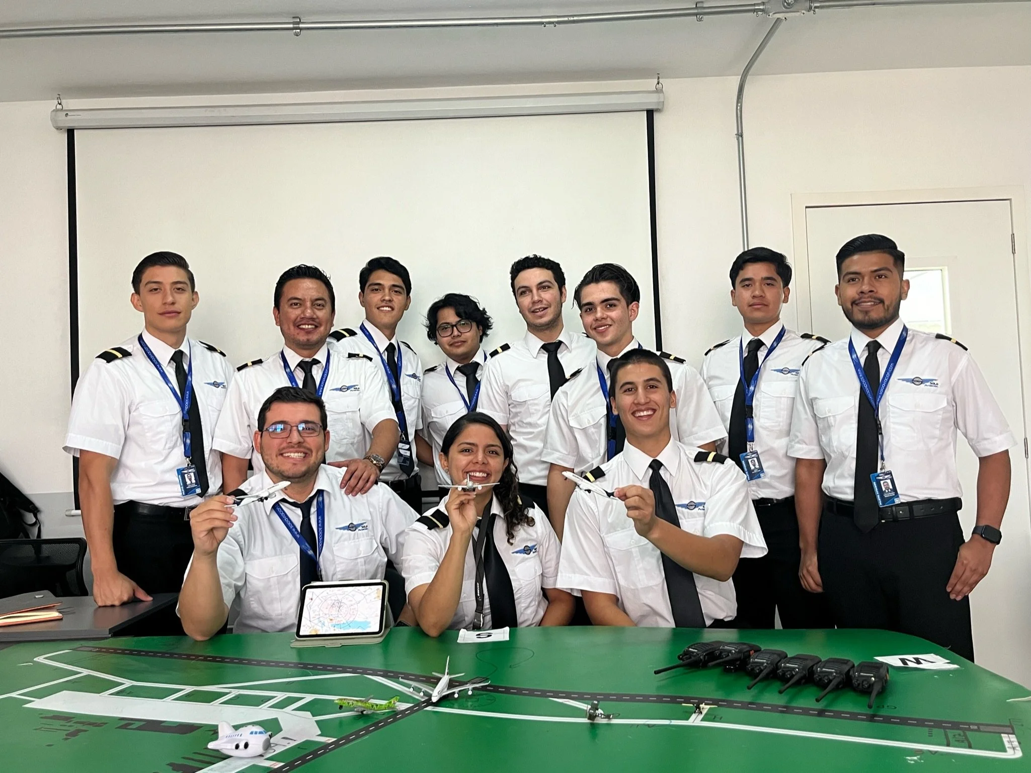 Grupo de personas en uniforme de pilotos en una sala de reuniones con mesa de simulador de aeropuerto, incluyendo aviones de juguete, radiocomunicadores y una tablet.
