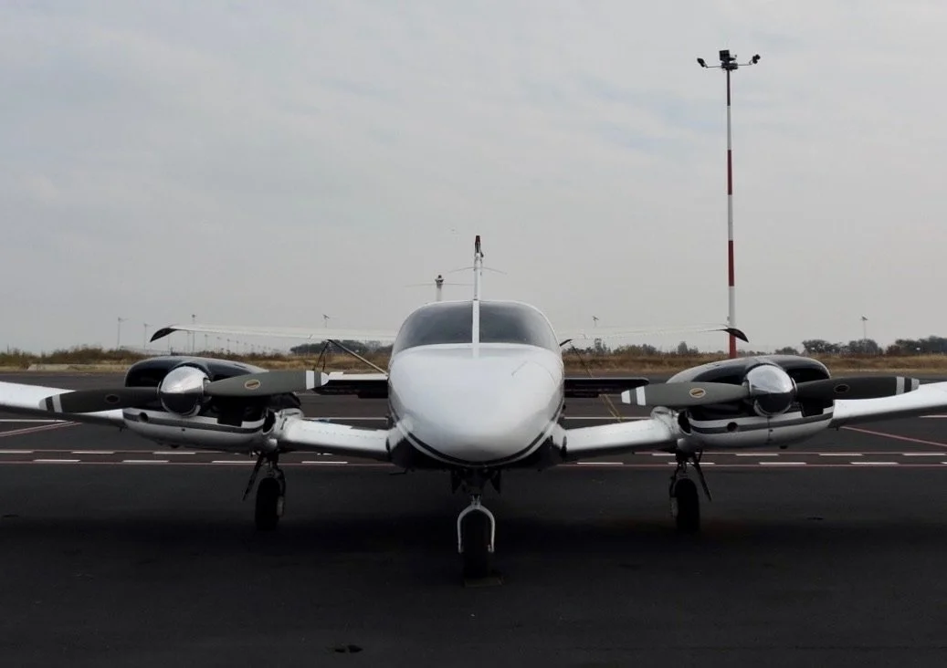 Avión pequeño en la pista del aeropuerto con torre de control y cielo nublado al fondo.