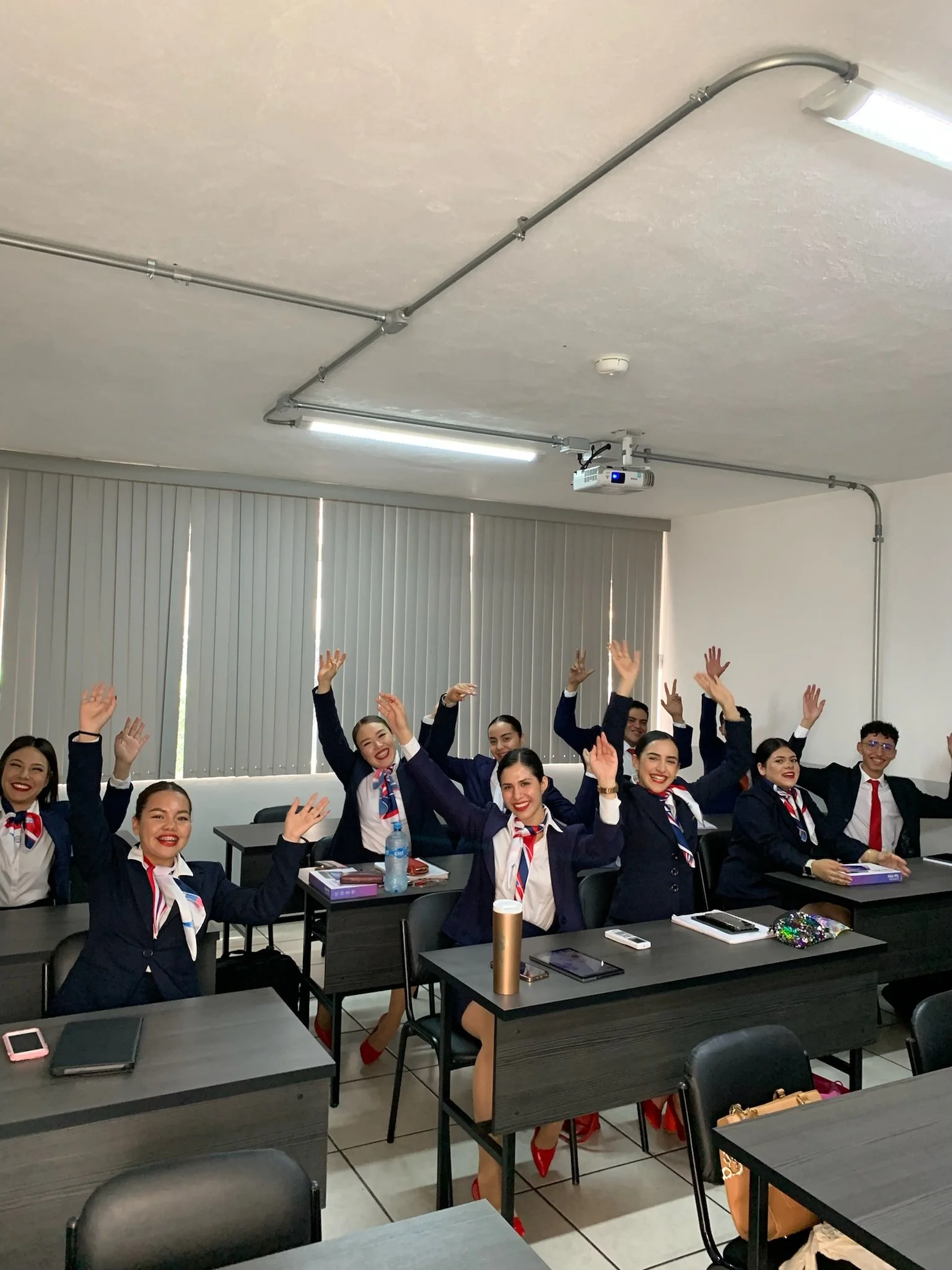 Grupo de estudiantes en uniforme con corbatas en clase, sonriendo y levantando las manos.