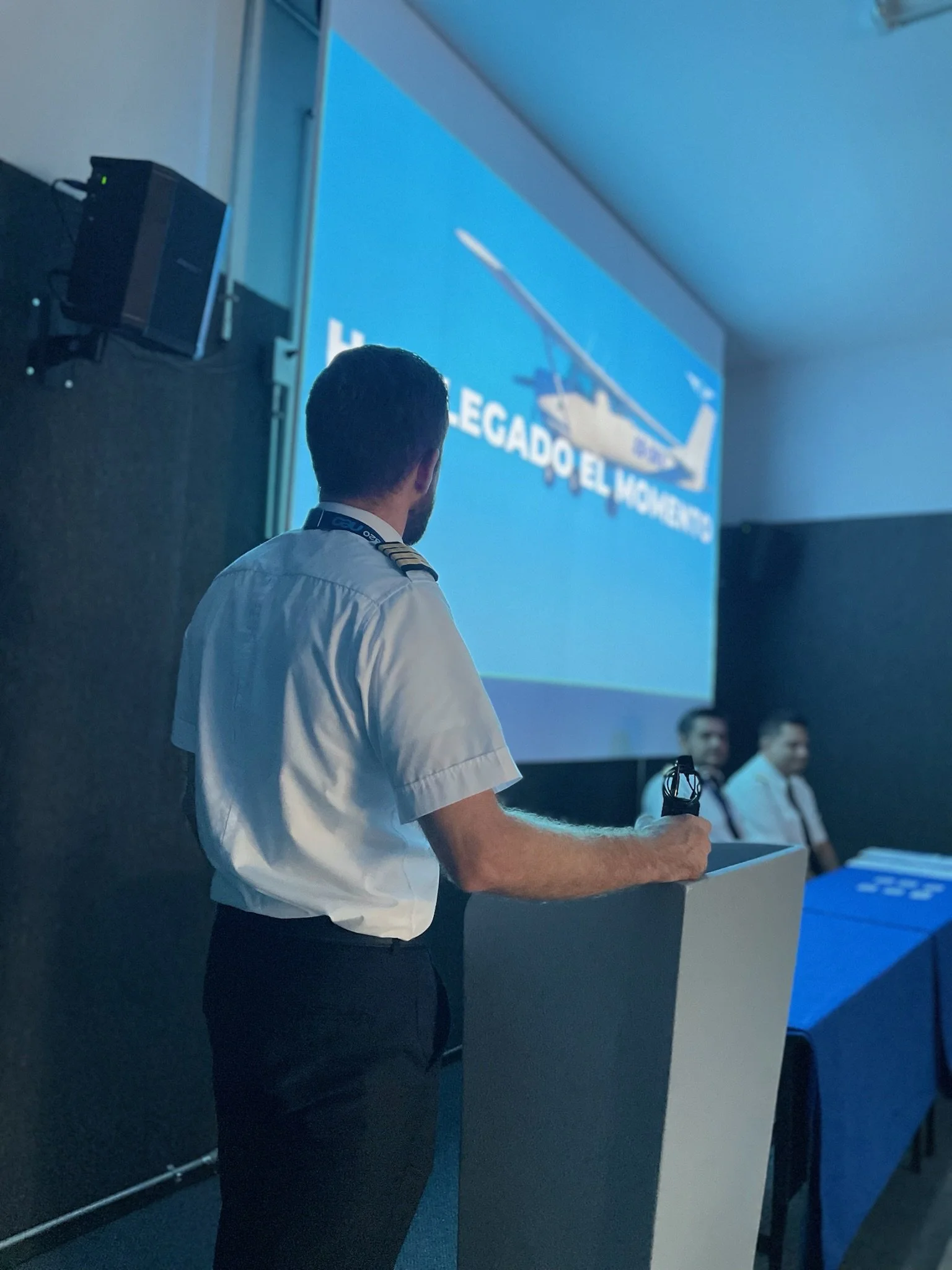 Hombre con uniforme militar de espaldas, dando una conferencia o presentación frente a una pantalla grande que muestra un helicóptero y texto en español. En la mesa a la derecha hay dos hombres con uniforme militar.