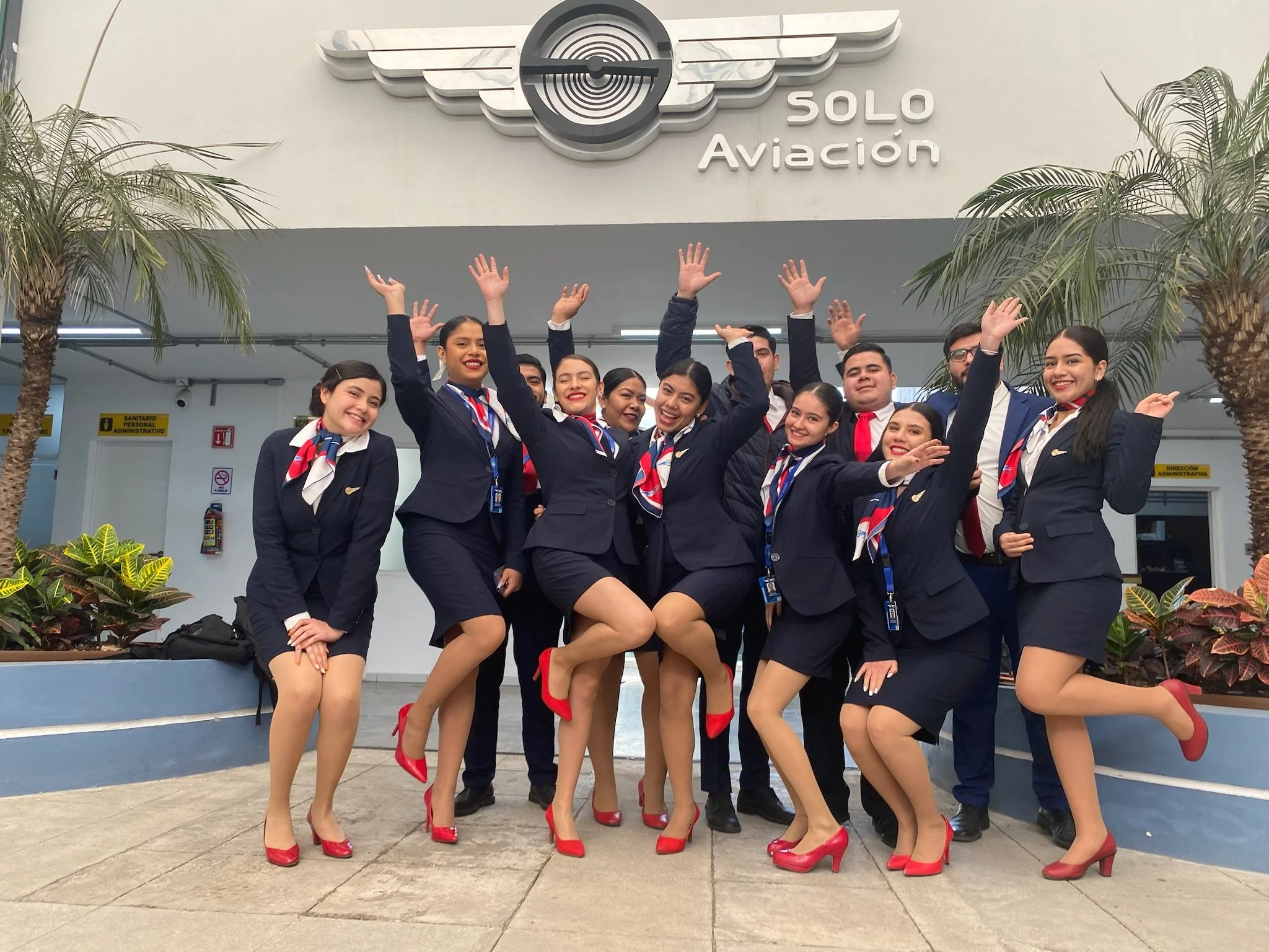 Grupo de empleados de aerolínea en uniforme en la entrada del aeropuerto Soló Aviación, celebrando con alegría.