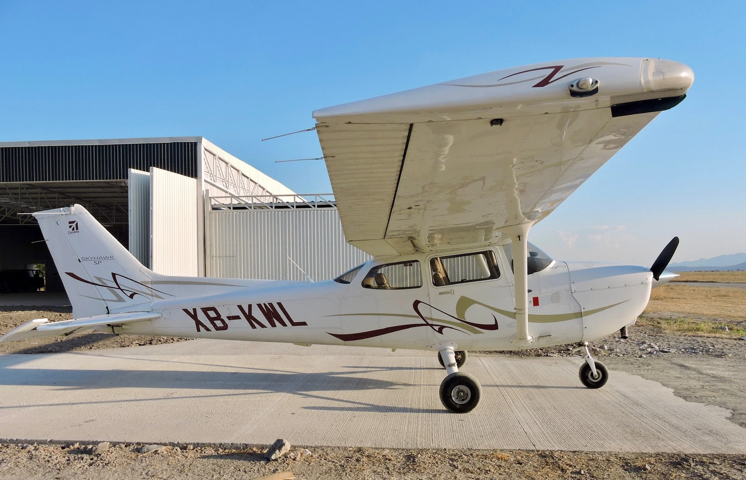 Avión pequeño con alas altas en una pista de aterrizaje con hangar al fondo, cielo despejado.