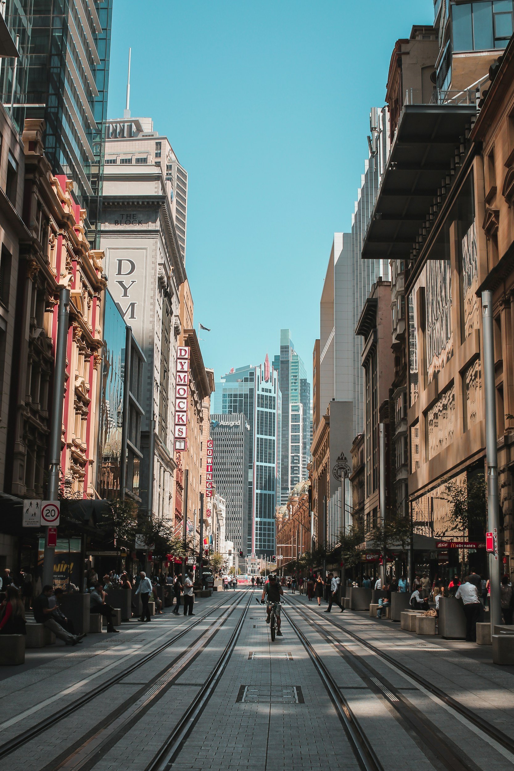 City street with tall modern buildings, people walking and cycling, tram tracks, and clear blue sky.